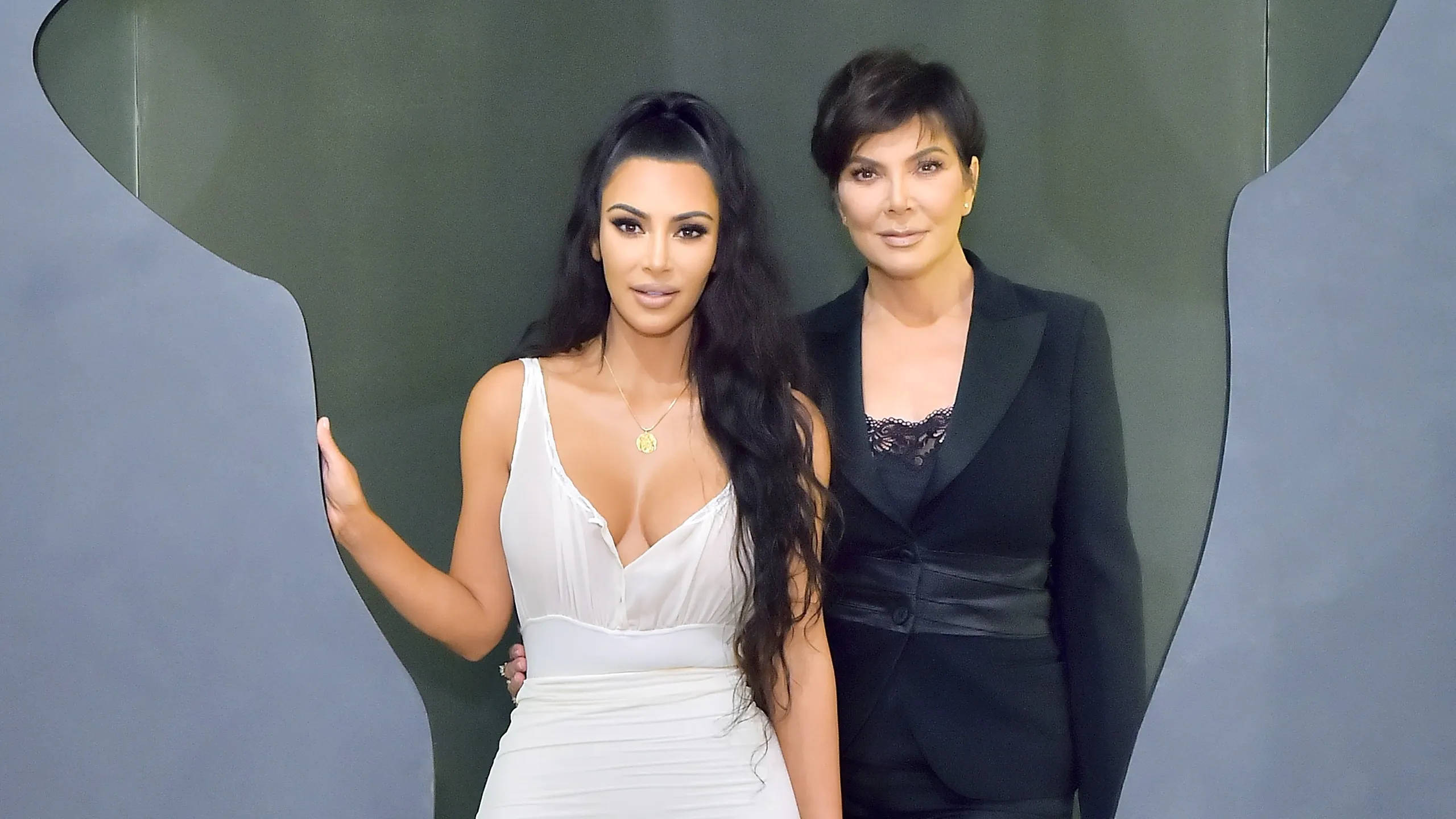kris 桌布 壁紙 — Kim Kardashian 和 Kris Jenner 穿著優雅服裝，在現代抽象灰色背景前自信地擺姿勢。