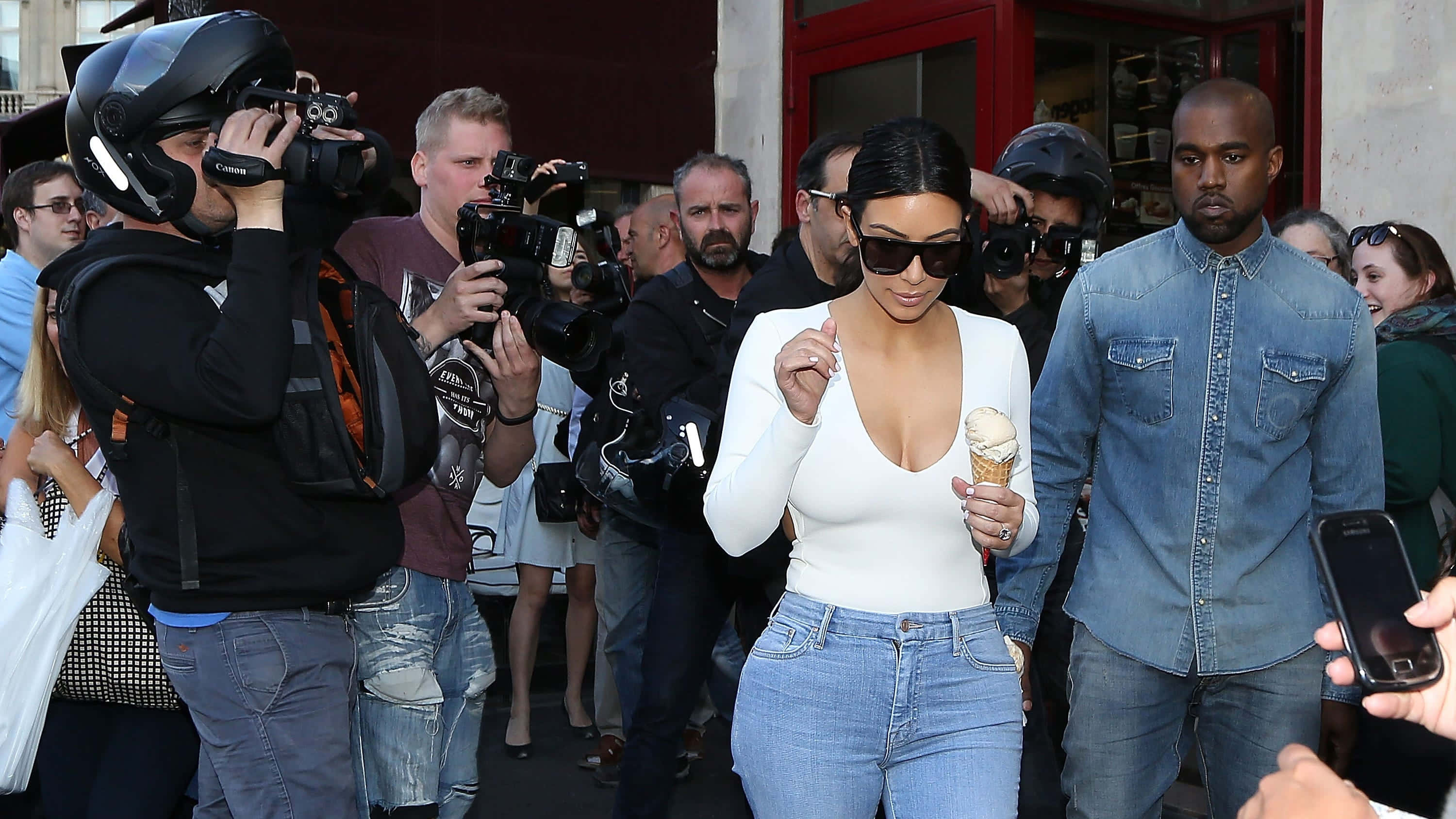 Paparazzi 桌布 壁紙 — Kim Kardashian 和 Kanye West 在拿著相機的狗仔隊人群中漫步，她一邊吃著冰淇淋。
