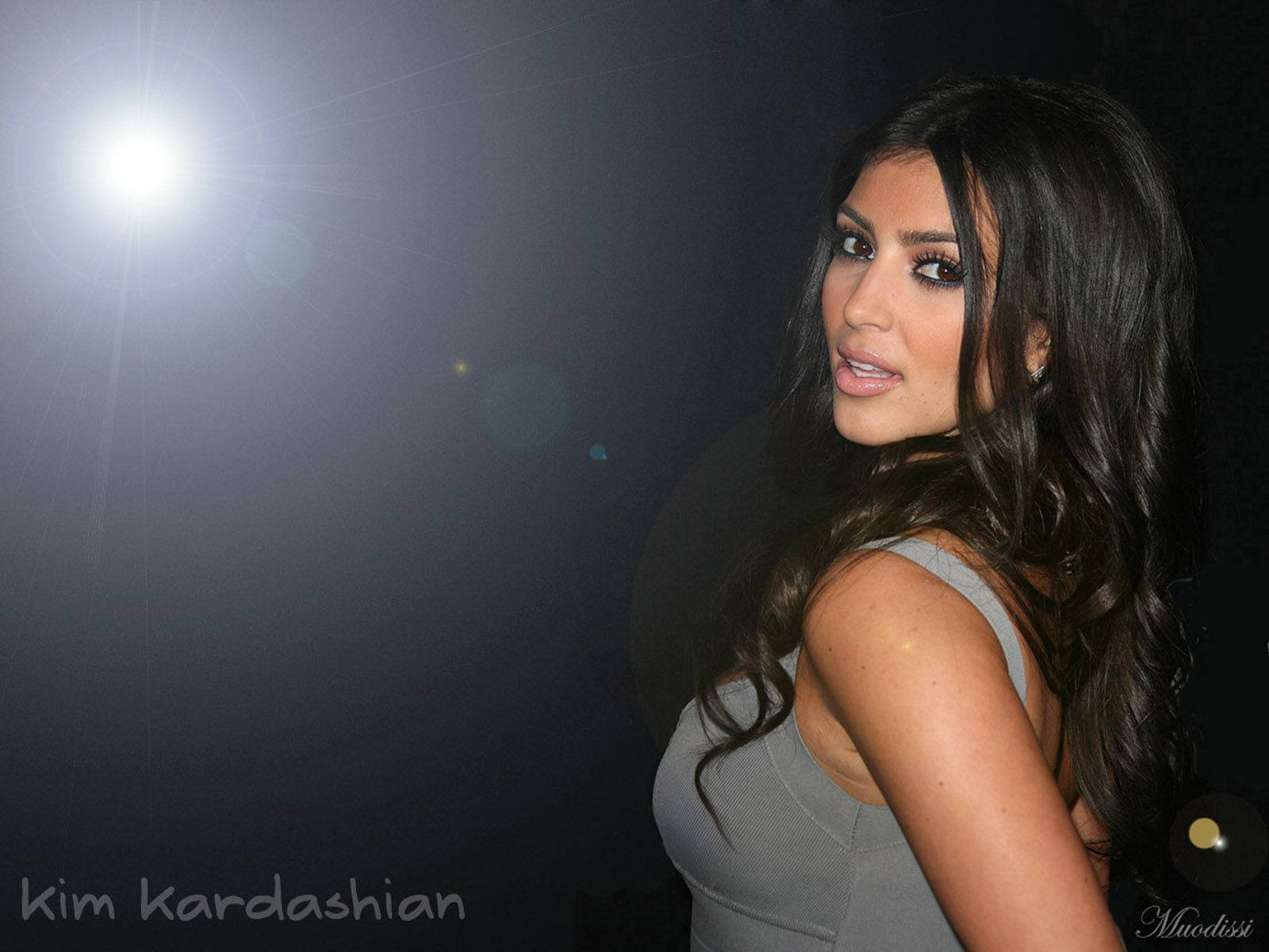 Kim Kardashian 桌布 壁紙 — Kim Kardashian 在黑暗背景下的明亮聚光燈下，穿著灰色上衣自信地擺姿勢，帶有鏡頭光暈。