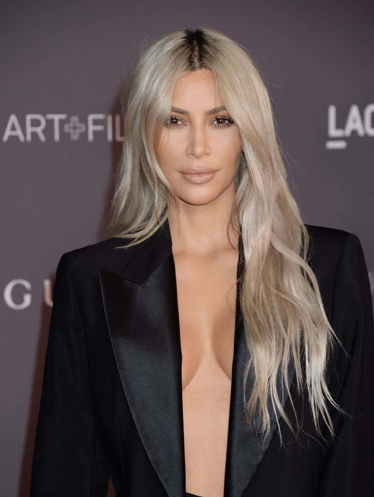 Kim Kardashian 桌布 壁紙 — Kim Kardashian 在品牌活動背景前，身穿時尚黑色西裝外套，留著長白金髮，擺出姿勢。