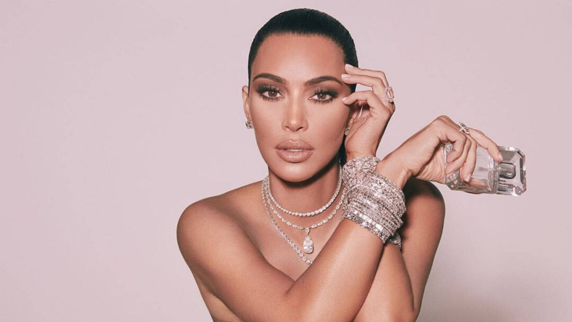 Kim Kardashian 桌布 壁紙 — Kim Kardashian 在柔和粉紅色背景的工作室自信地擺姿勢，手持一個飾有閃亮珠寶的香水瓶。