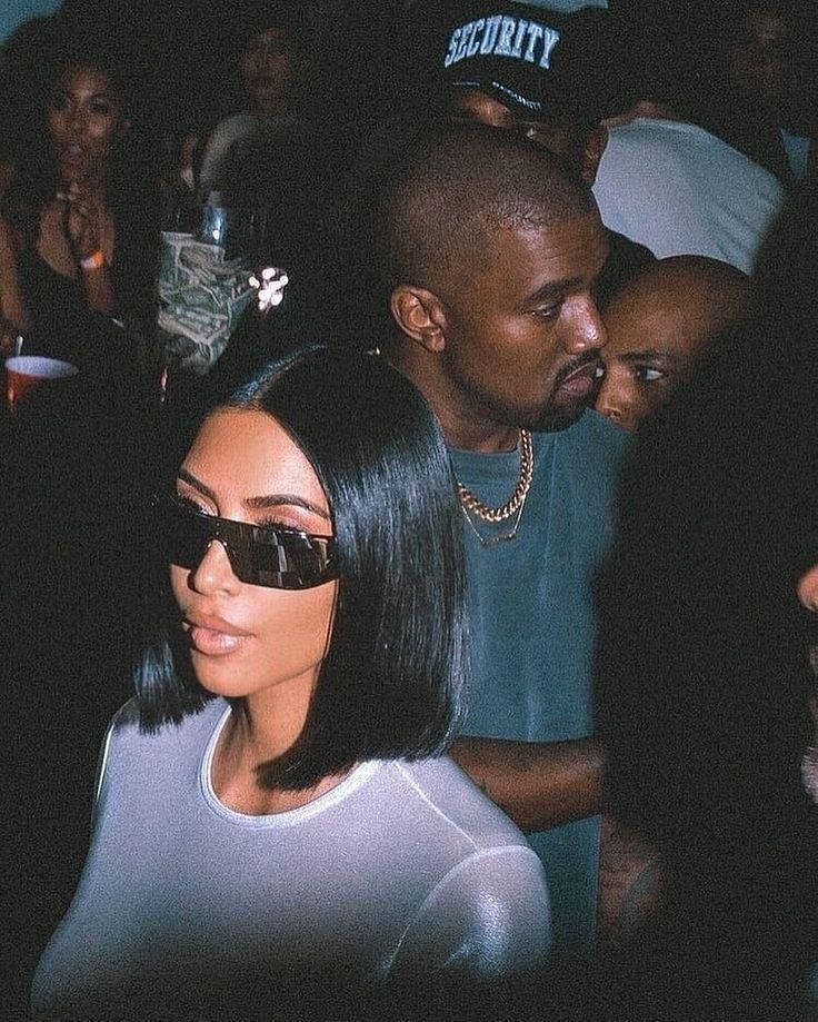 Kim Kardashian 桌布 壁紙 — Kim Kardashian 戴著太陽眼鏡，身穿透視上衣，站在燈光昏暗、擠滿人的派對中，Kanye West 在她身後。