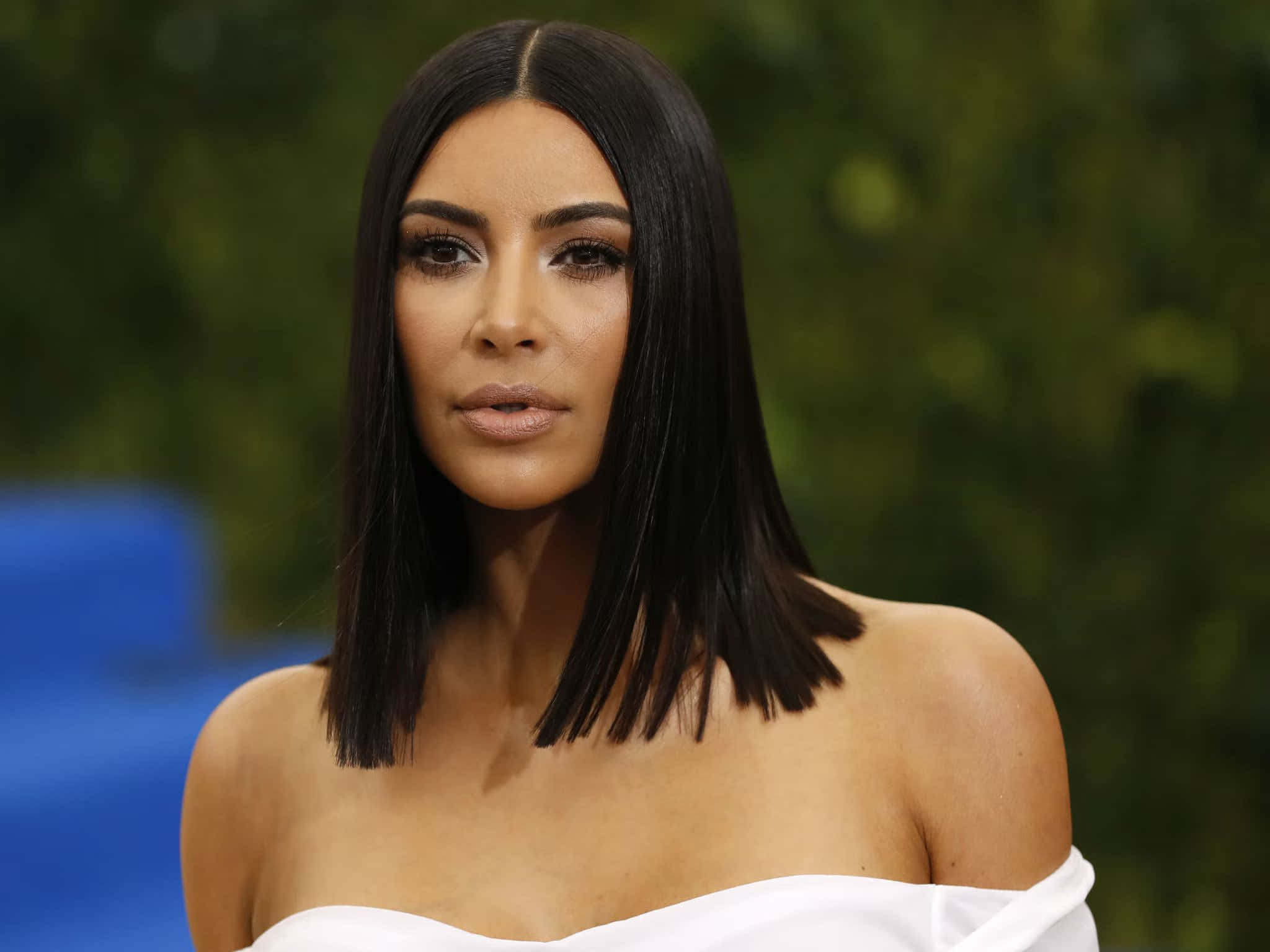 Kim Kardashian 桌布 壁紙 — Kim Kardashian 穿著白色露肩連衣裙，搭配光滑的黑色鮑伯頭，在模糊的綠色和藍色背景前擺姿勢。