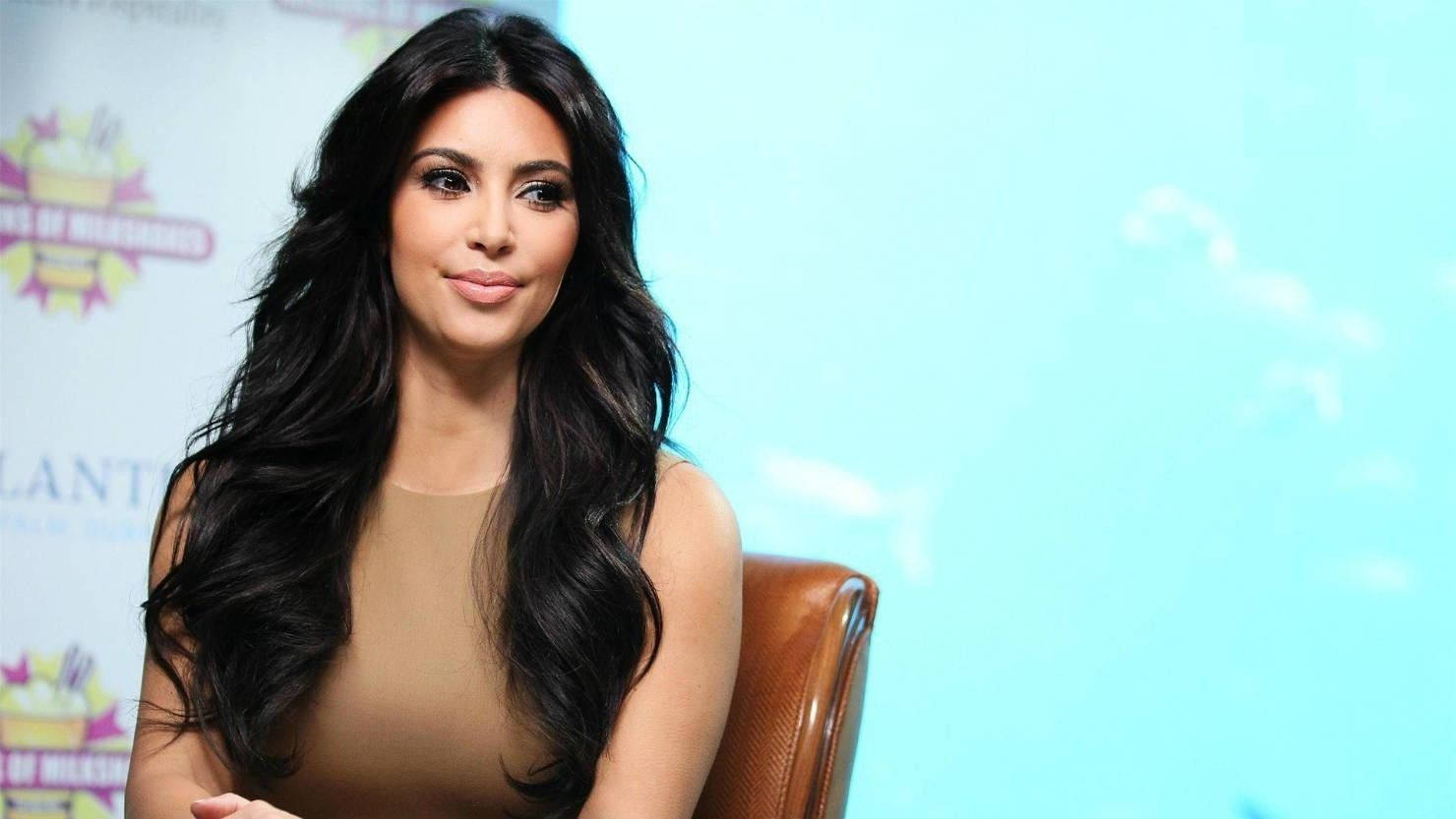 Kim Kardashian 桌布 壁紙 — Kim Kardashian 端坐在棕色皮椅上，背景是明亮的藍綠色，帶有活動品牌標識。