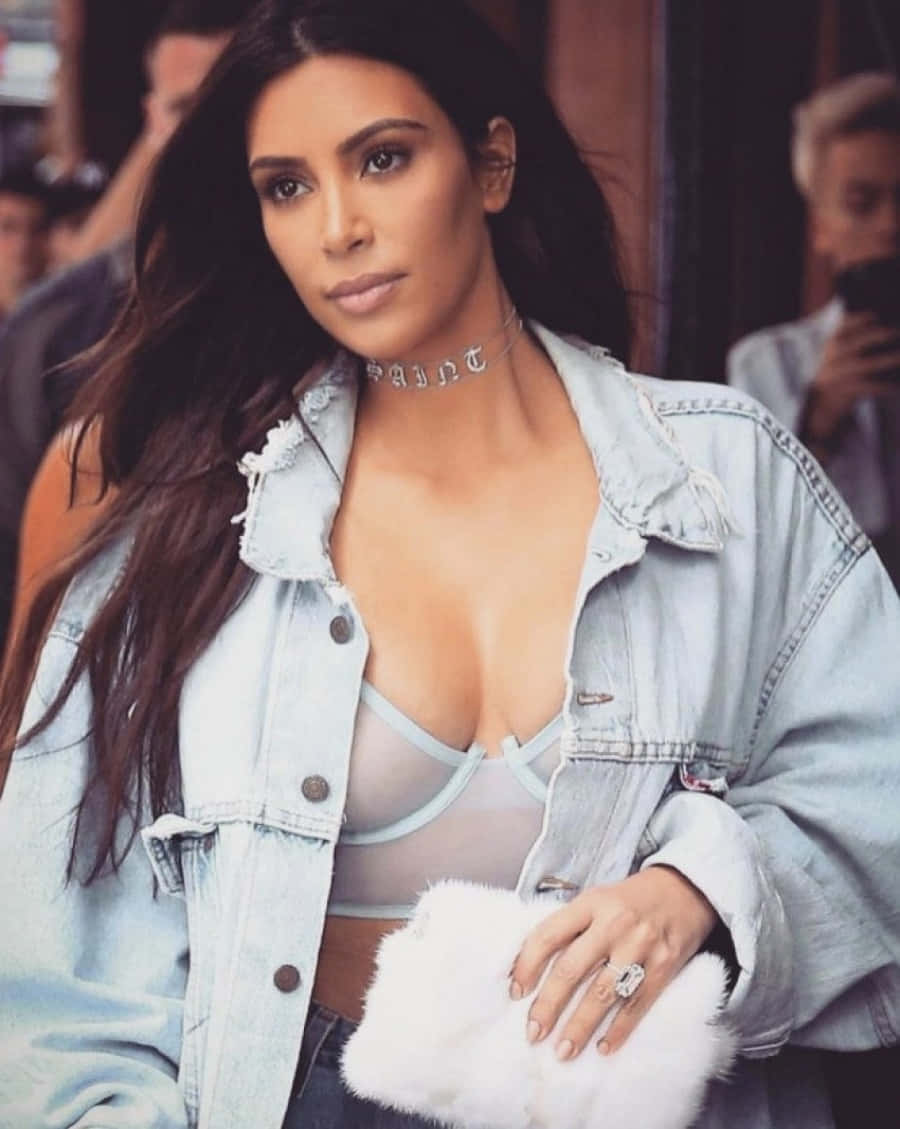 Kim Kardashian 桌布 壁紙 — Kim Kardashian 自信地擺姿勢，身穿淺色牛仔外套內搭透視上衣，手持白色毛絨手拿包。