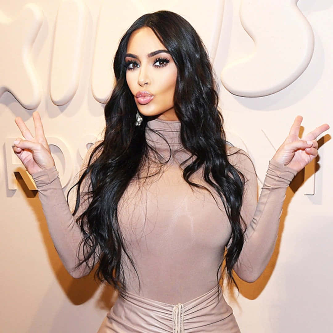 Kim Kardashian 桌布 壁紙 — Kim Kardashian 自信地擺姿勢，身穿透明米色連體衣，對著白色品牌牆，雙手比出和平手勢。