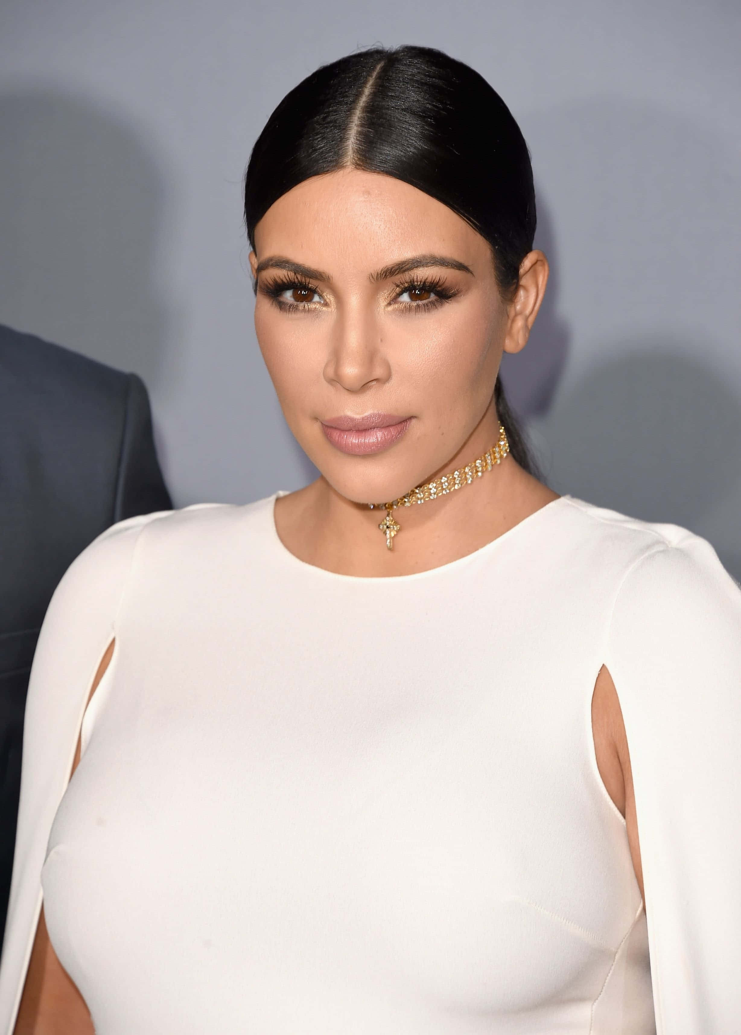 Kim Kardashian 桌布 壁紙 — Kim Kardashian 身穿一襲帶有鏤空設計的修身白色連衣裙，搭配金色頸鍊，在柔和的灰色背景前擺姿勢。