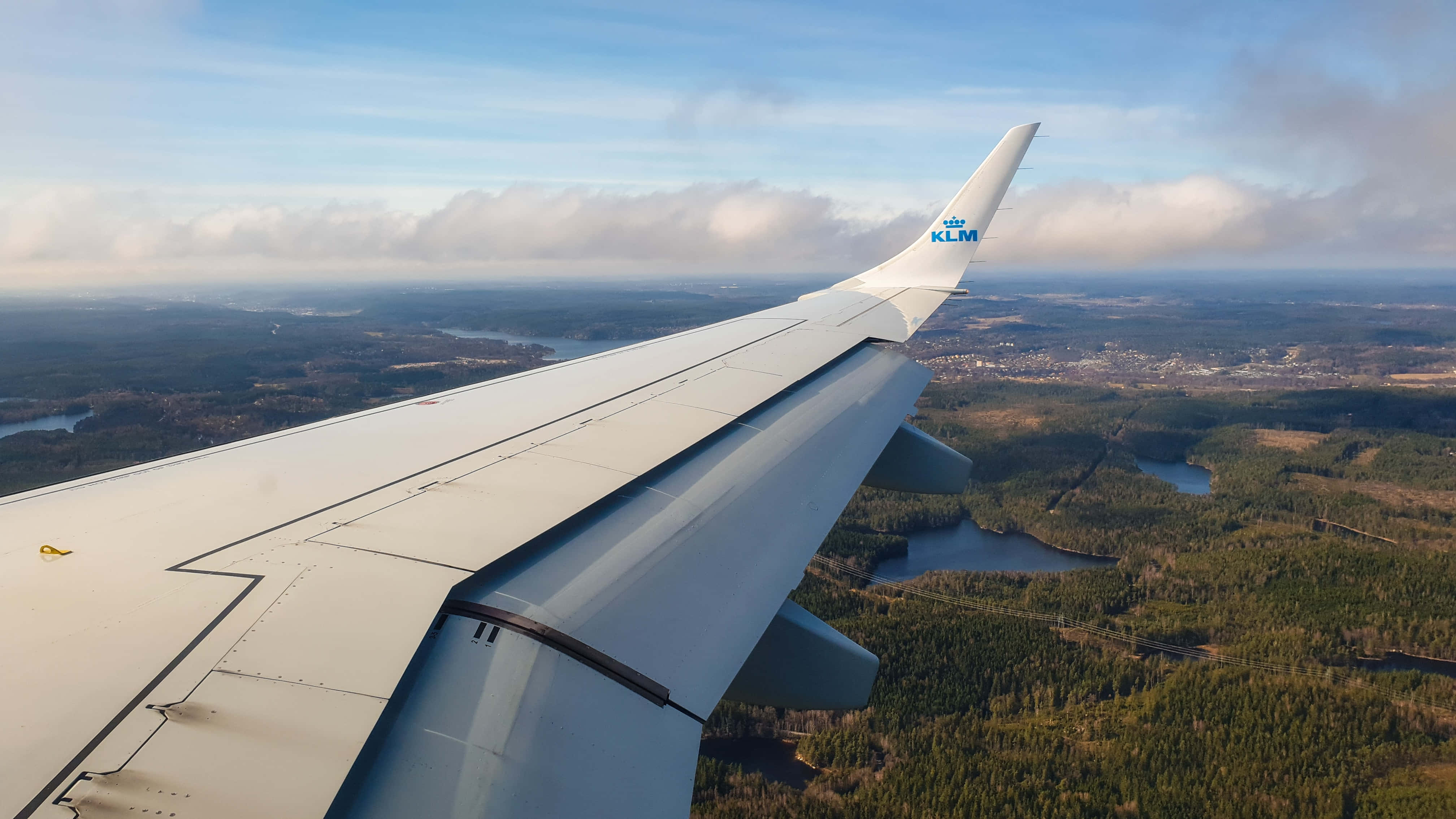 飛機 桌布 壁紙 — KLM 飛機的機翼延伸在森林、湖泊和城鎮的景觀之上，天空部分多雲。