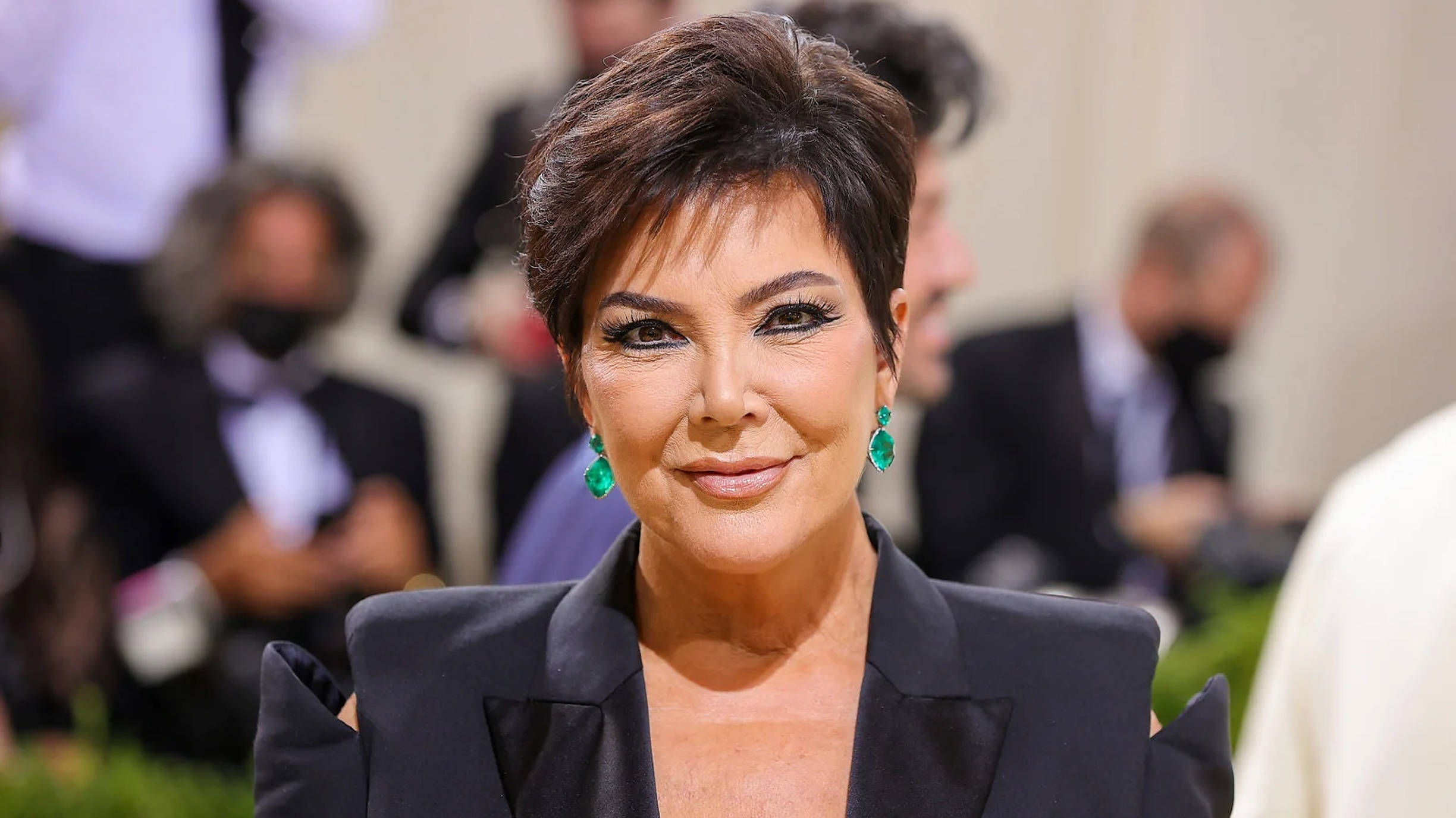 kris 桌布 壁紙 — Kris Jenner 在一個華麗的活動中，自信地微笑，身穿黑色西裝外套，搭配搶眼的綠色耳環。