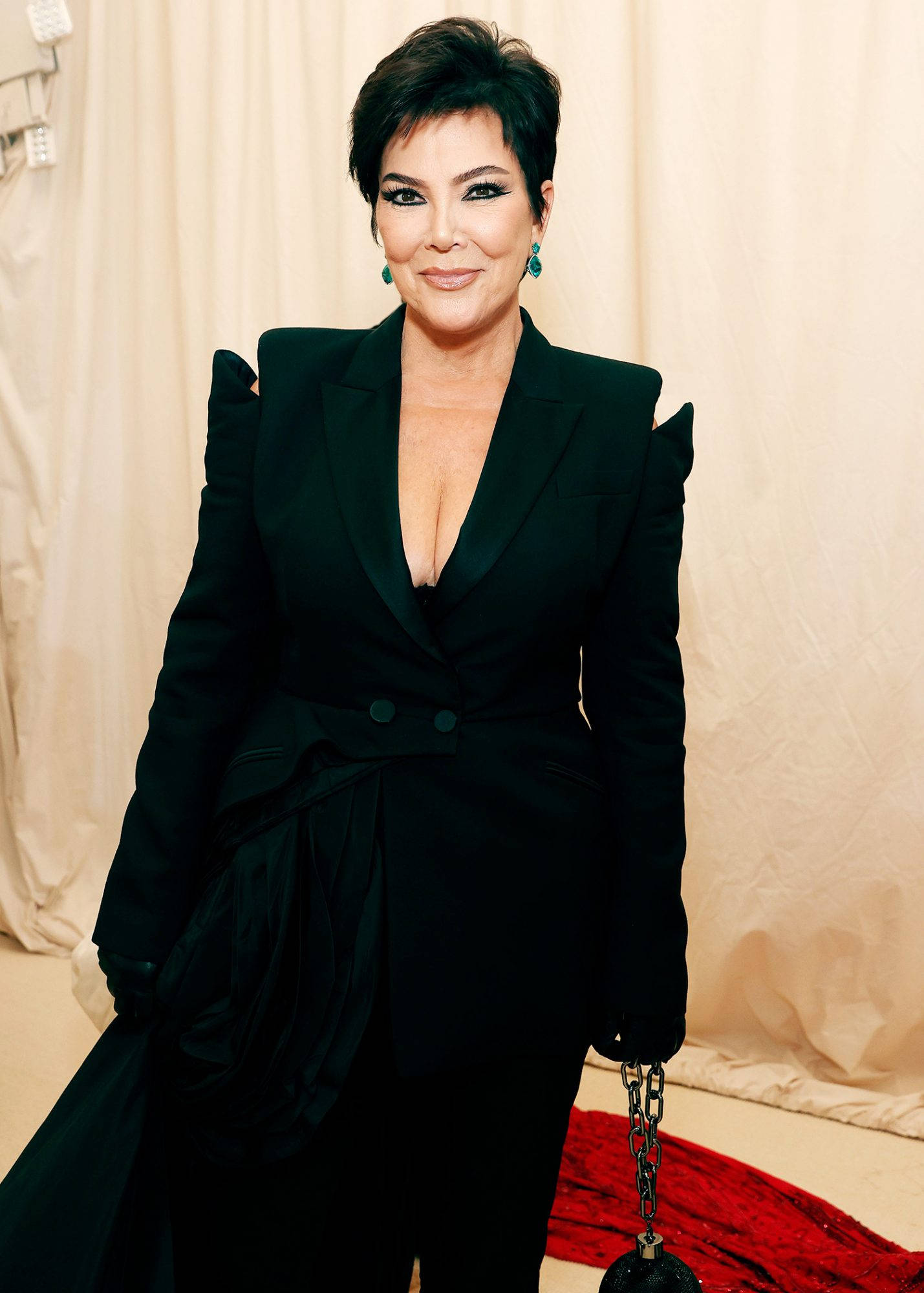 kris 桌布 壁紙 — Kris Jenner 在奶油色背景前自信地擺姿勢，身穿一件帶有荷葉邊肩部和鏈條流蘇手袋的戲劇性黑色西裝外套。