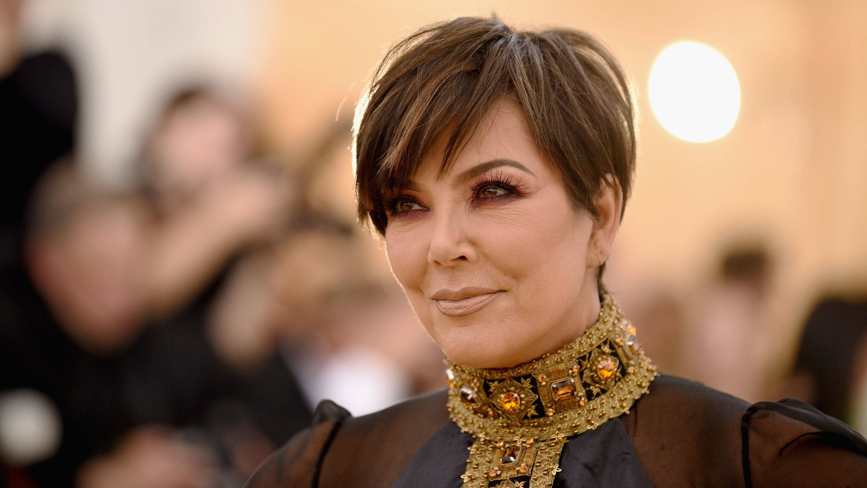 kris 桌布 壁紙 — Kris Jenner 在紅毯活動中自信地微笑，戴著華麗的金色珠寶頸鍊，身後是模糊的人群。