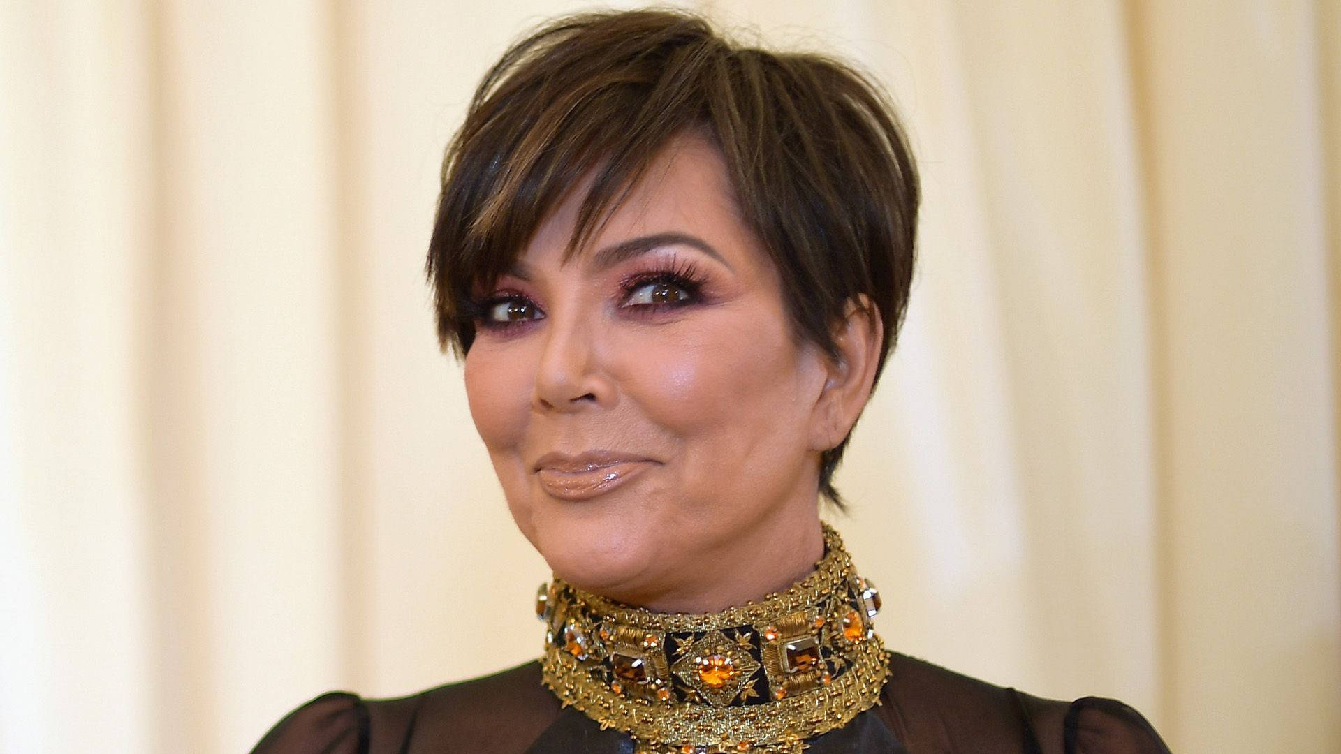 kris 桌布 壁紙 — Kris Jenner 戴著一條鑲有寶石的金色頸鍊，在柔和的奶油色窗簾背景下溫暖地微笑。