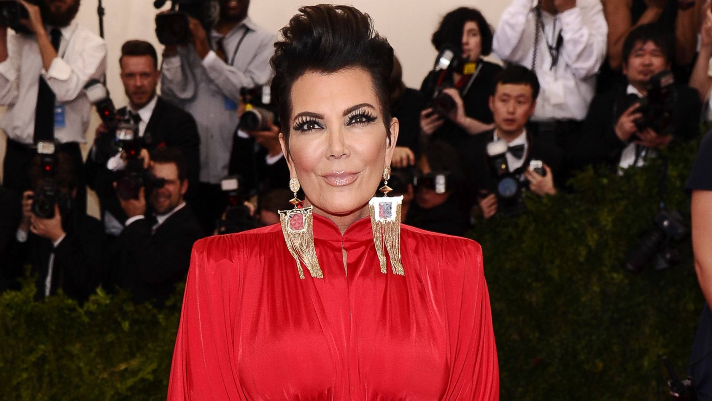 kris 桌布 壁紙 — Kris Jenner 穿著大膽的紅色緞面禮服和超大金色耳環，在華麗的紅地毯活動中自信地擺姿勢。