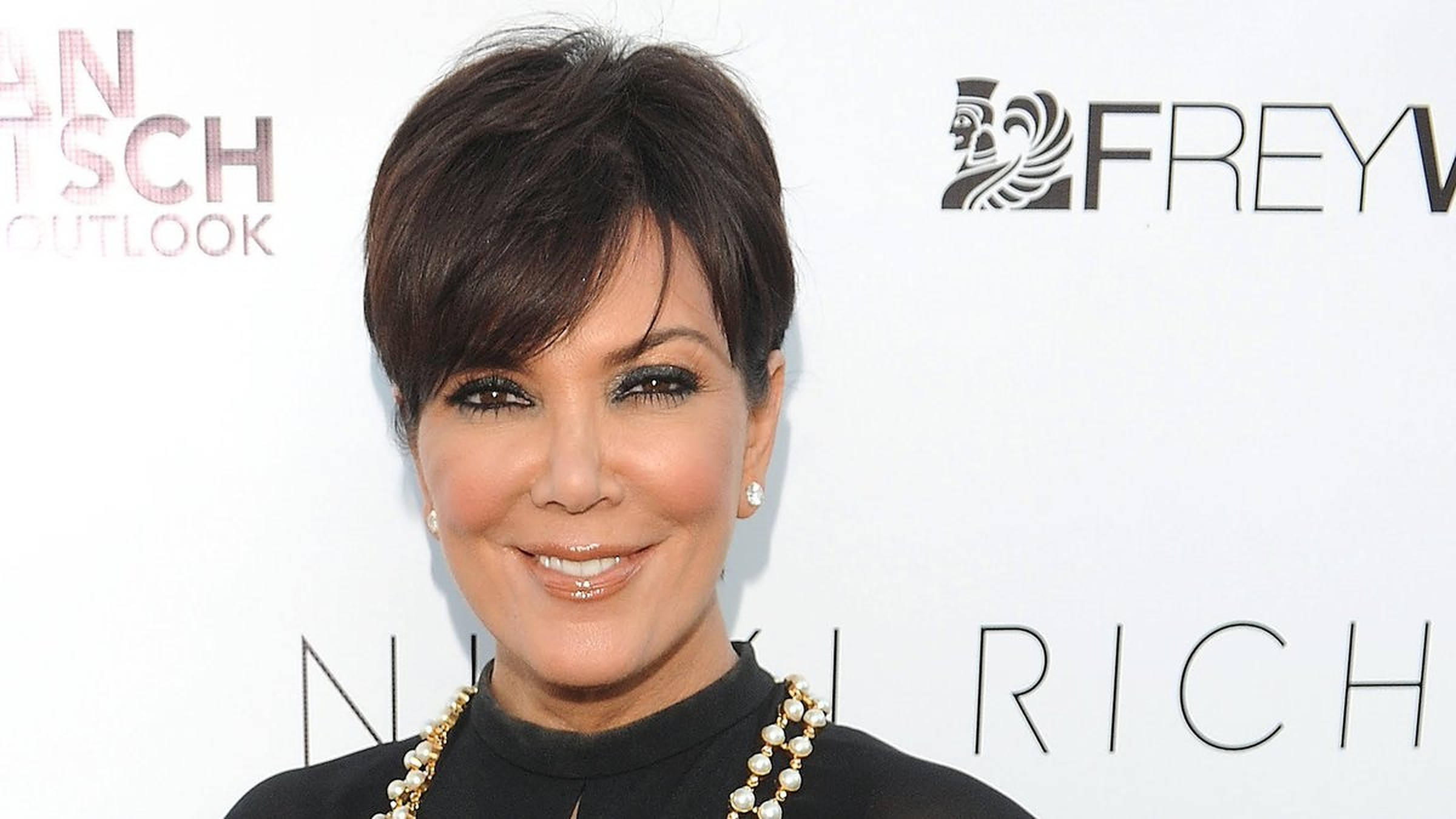 kris 桌布 壁紙 — Kris Jenner 穿著黑色上衣和珍珠項鍊，在品牌活動背景下對著鏡頭溫暖微笑。
