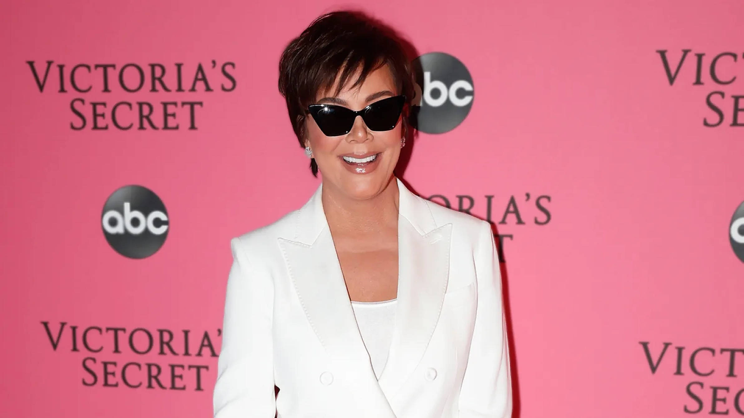 kris 桌布 壁紙 — Kris Jenner 身穿白色西裝外套和黑色太陽眼鏡，在粉紅色Victoria's Secret品牌背景前自信地微笑。