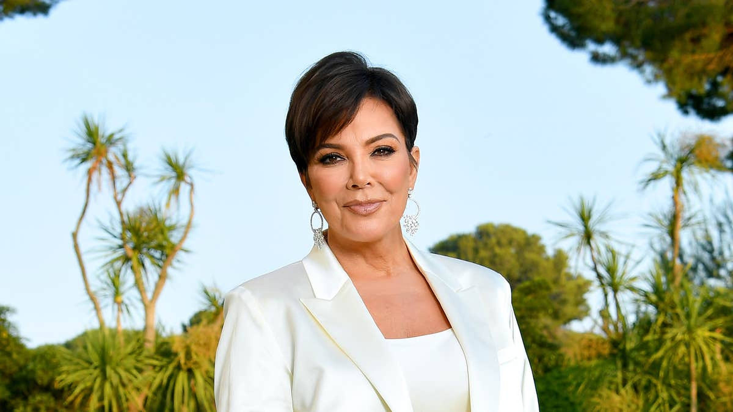 kris 桌布 壁紙 — Kris Jenner 身穿白色西裝外套，在棕櫚樹和晴朗藍天的背景下自信地微笑。