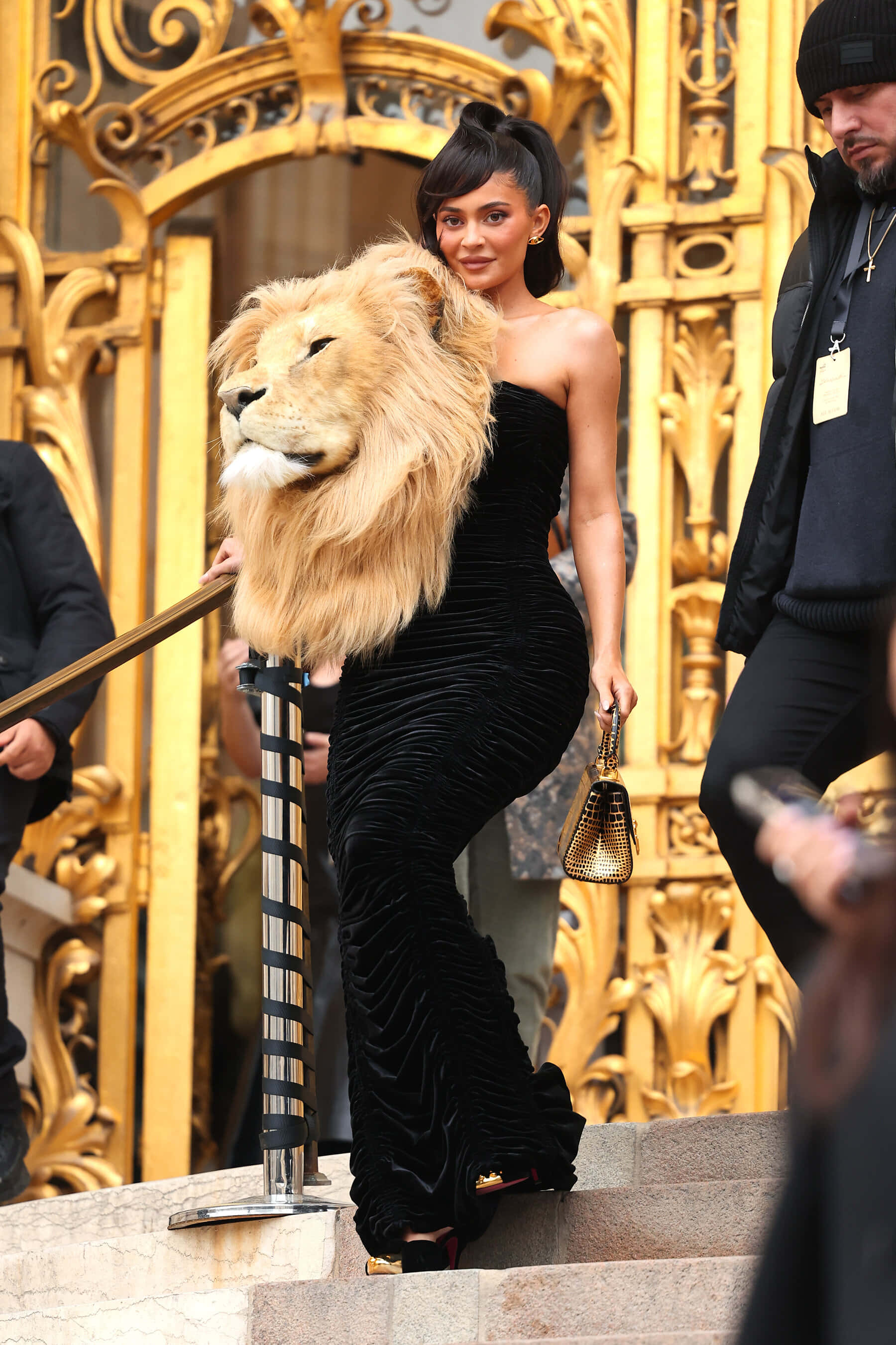 Lion Head 桌布 壁紙 — Kylie Jenner 身穿黑色天鵝絨長裙，在華麗的金色大門旁，伴隨著獅頭道具走下樓梯。