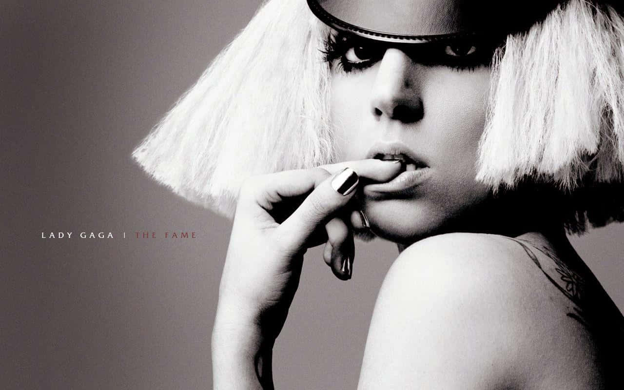女神卡卡 桌布 壁紙 — Lady Gaga 以單色調呈現，戴著白色鮑伯頭假髮和深色太陽眼鏡，手指靠近嘴唇，肩膀上的刺青清晰可見。