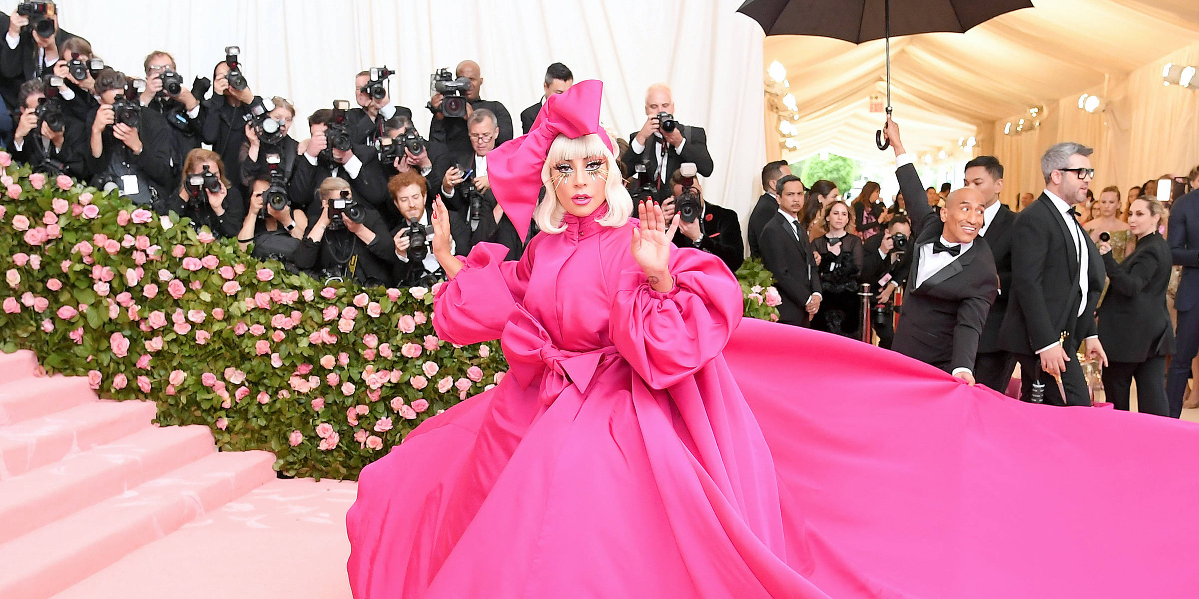 Gala 桌布 壁紙 — Lady Gaga 身穿大膽的粉紅色長袍，在 Met Gala 上向攝影師揮手，身後有花卉樓梯和人群。