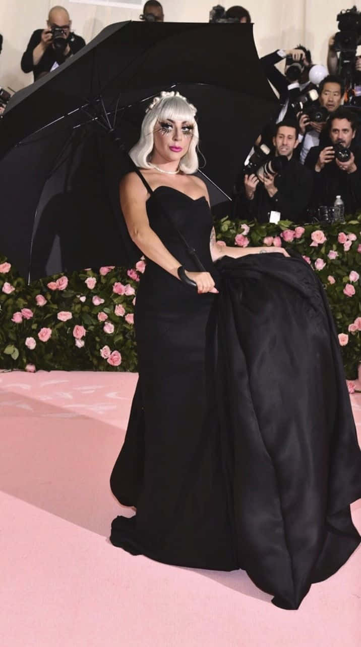 Met Gala 桌布 壁紙 — Lady Gaga 身穿戲劇性的黑色長裙，在粉紅色地毯上拿著一把大黑傘，周圍是攝影師和粉紅玫瑰。