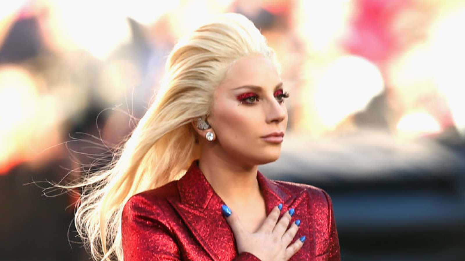 女神卡卡 桌布 壁紙 — Lady Gaga 身穿閃亮紅色外套，手放胸前，金髮飄逸，大膽紅色眼妝，背景模糊火焰。