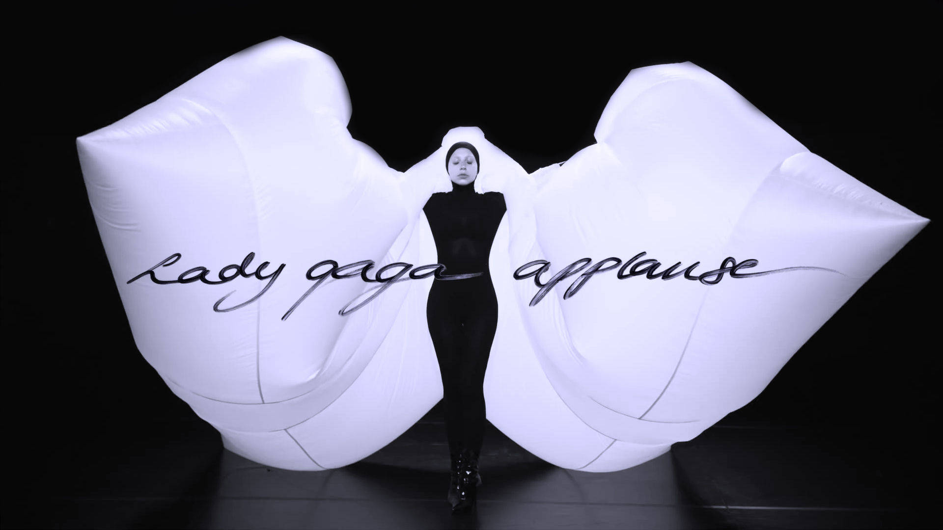 lady 桌布 壁紙 — Lady Gaga 身穿黑色緊身連衣褲站在舞台中央，兩側有巨大的白色充氣手，以草書拼出她的名字。