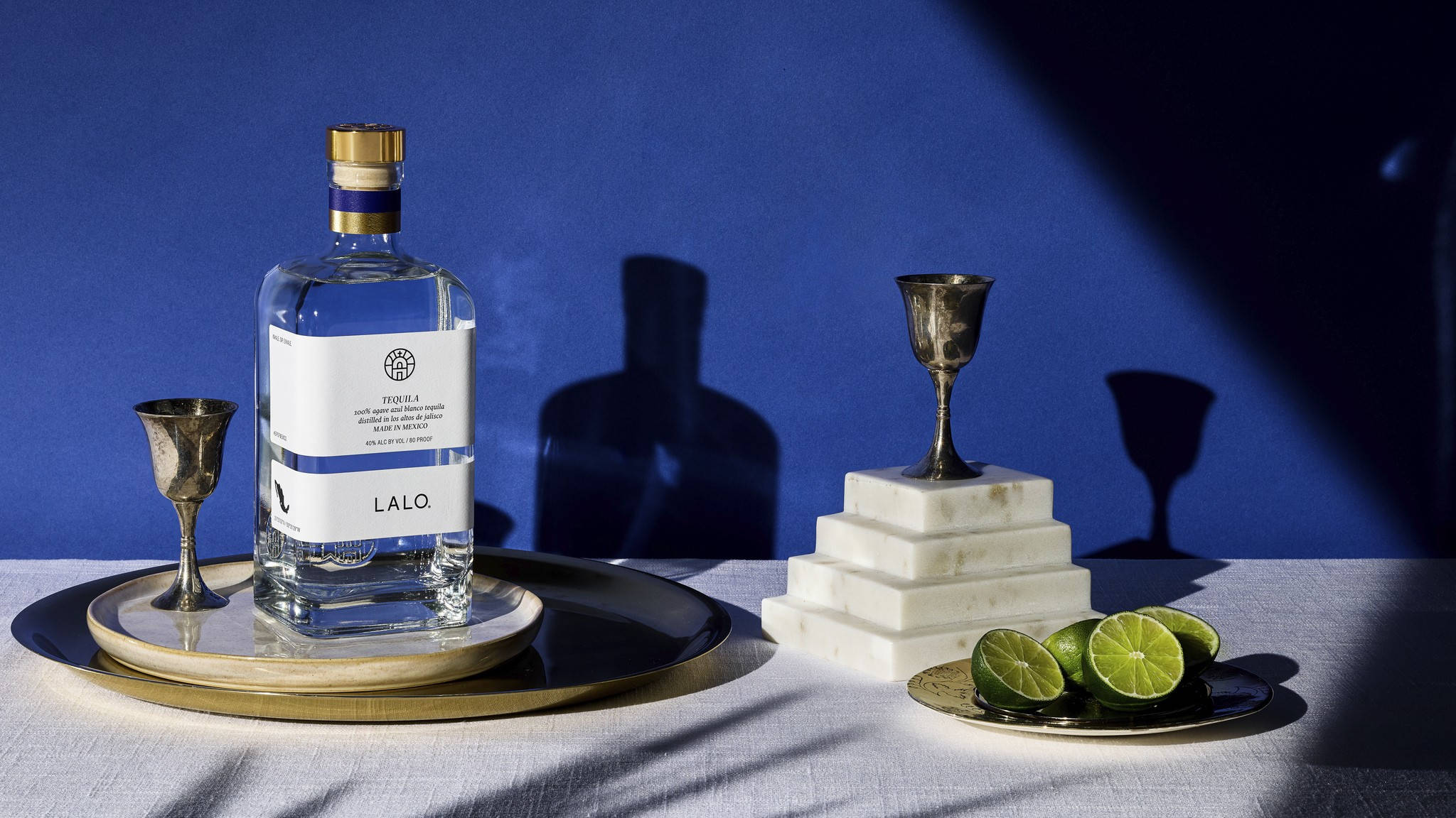 alcohol 桌布 壁紙 — LALQ 龍舌蘭酒的時尚瓶子放在金色托盤上，旁邊有銀色高腳杯和切片萊姆，背景是深藍色，帶有戲劇性的陰影。