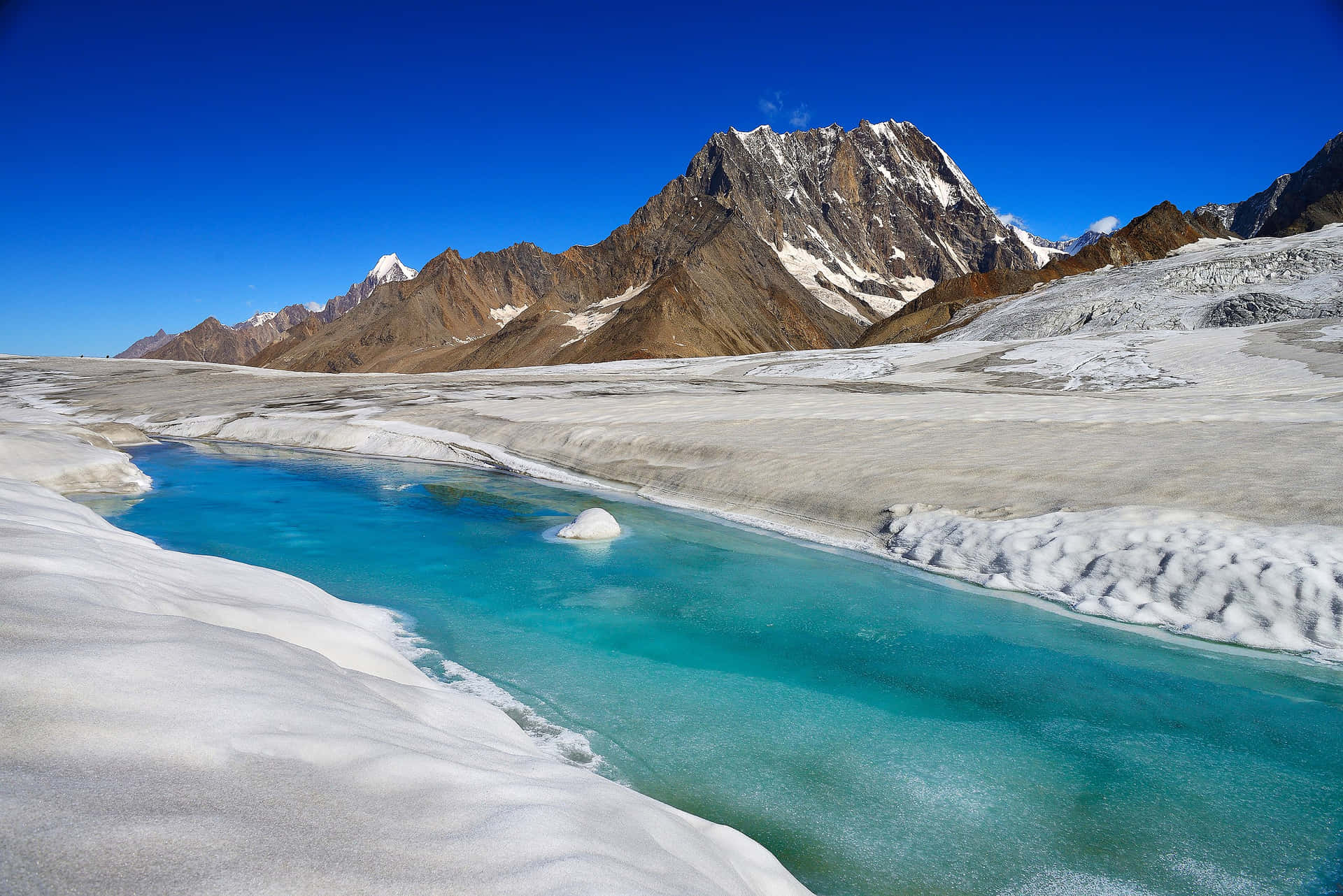  壁紙 — 一條充滿活力的藍綠色冰川河流蜿蜒穿過雪原，在清澈的藍天下聳立著岩石山峰。