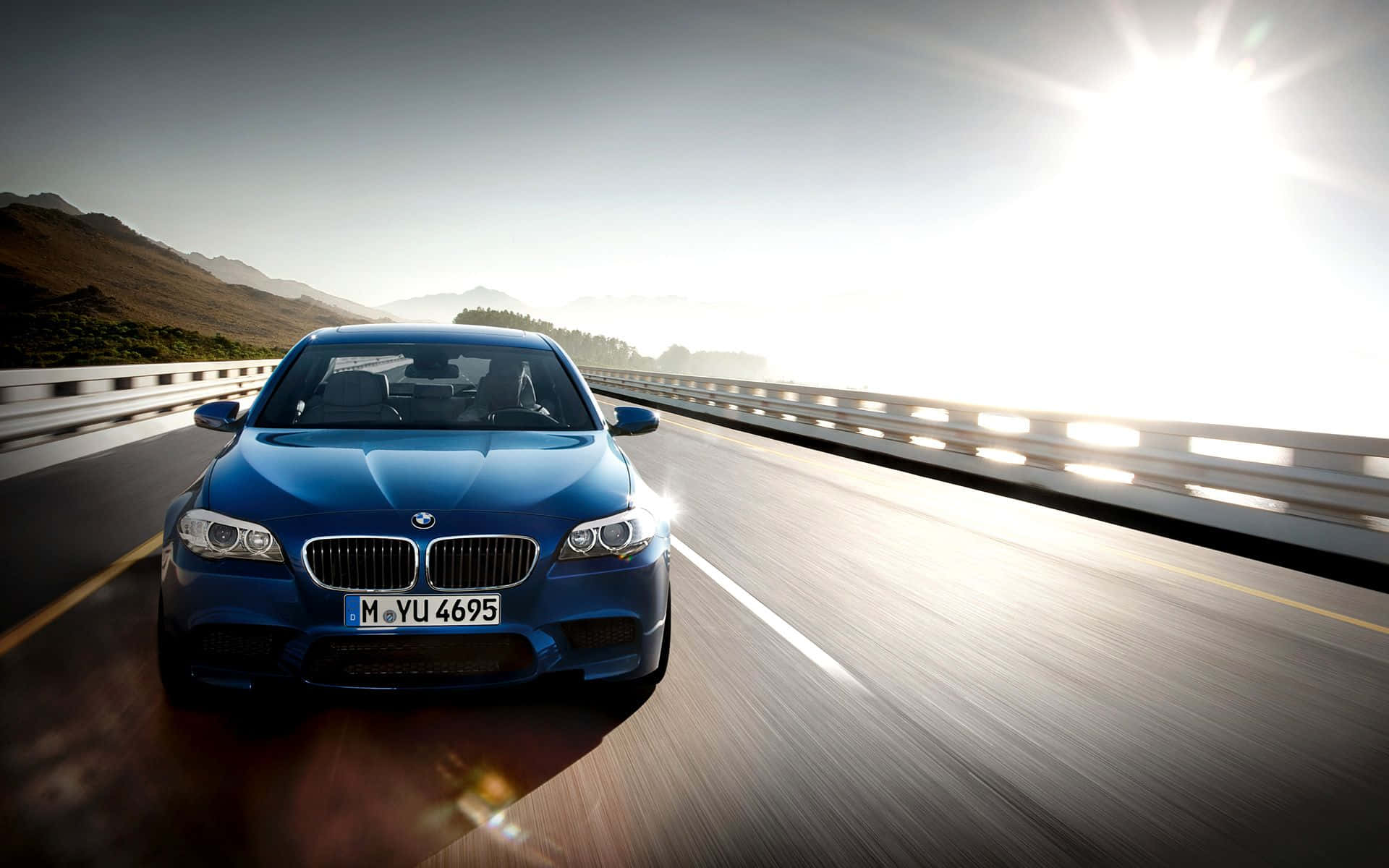 Bmw M5 桌布 壁紙 — 一輛充滿活力的藍色BMW跑車，在陽光普照的山區高速公路上奔馳，帶有動態模糊和鏡頭光暈效果。