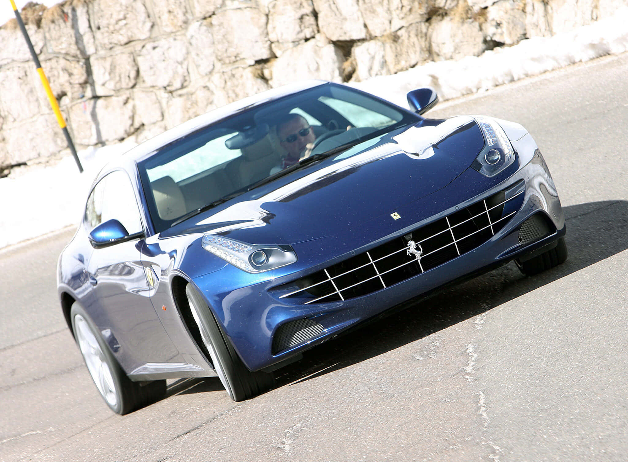 Ferrari Ff 桌布 壁紙 — 一架流線型藍色法拉利跑車沿著陽光明媚的山路，在風化的石牆旁行駛。
