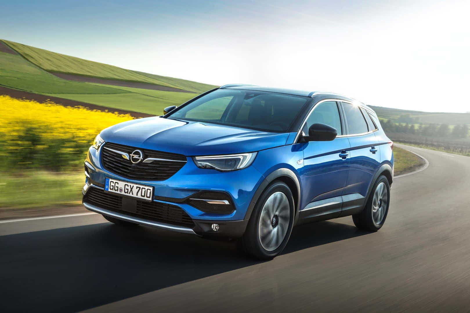 Opel Grandland X 桌布 壁紙 — 一輛充滿活力的藍色Opel SUV沿著蜿蜒的道路穿過起伏的綠色山丘和明亮的黃色花田飛馳。