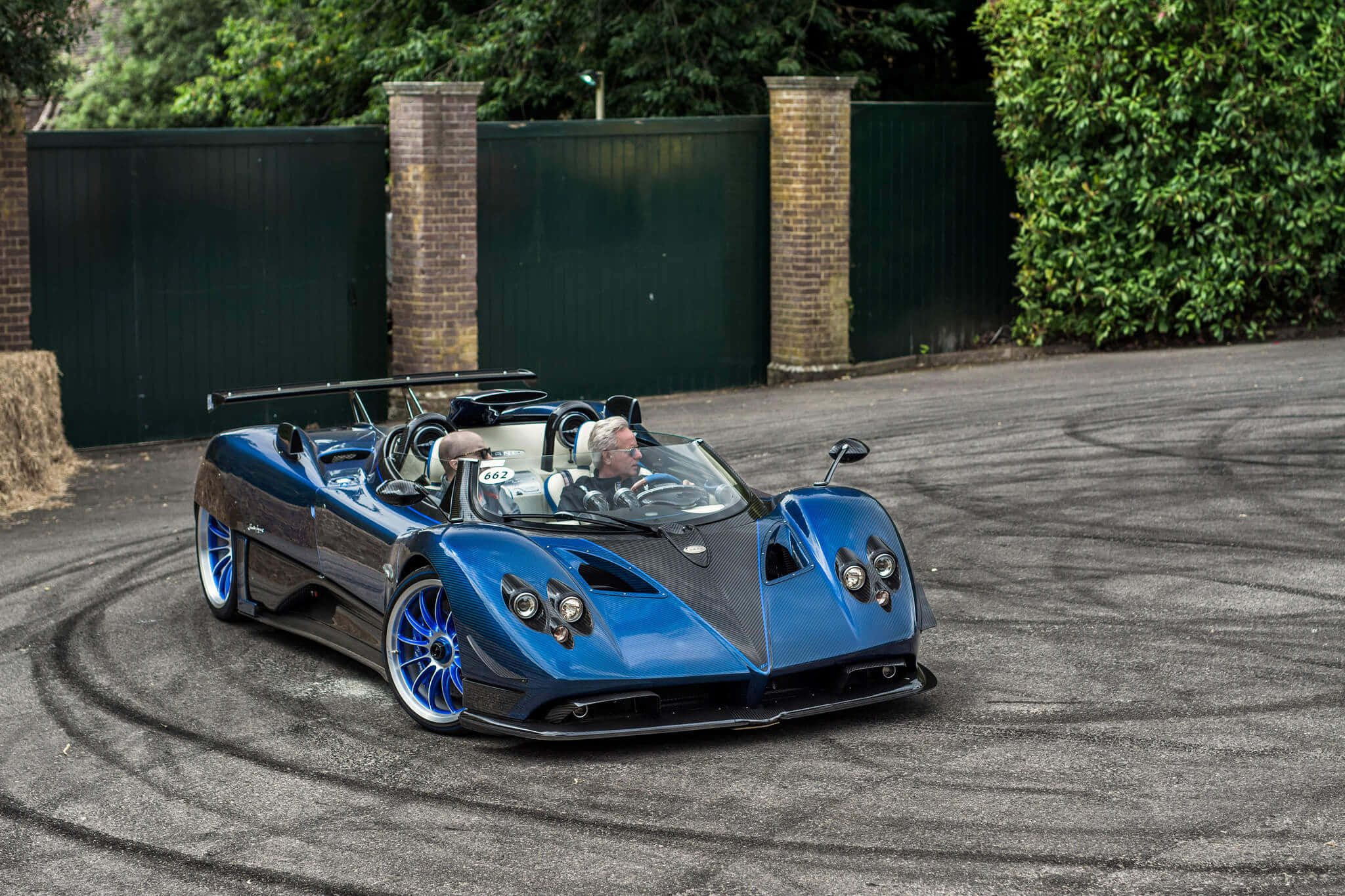 Pagani Zonda Hp Barchetta 桌布 壁紙 — 一輛搶眼的藍色Pagani Zonda跑車在司機駕駛下，於鋪砌的車道上進行飄移，車道兩旁有綠色籬笆和磚柱。