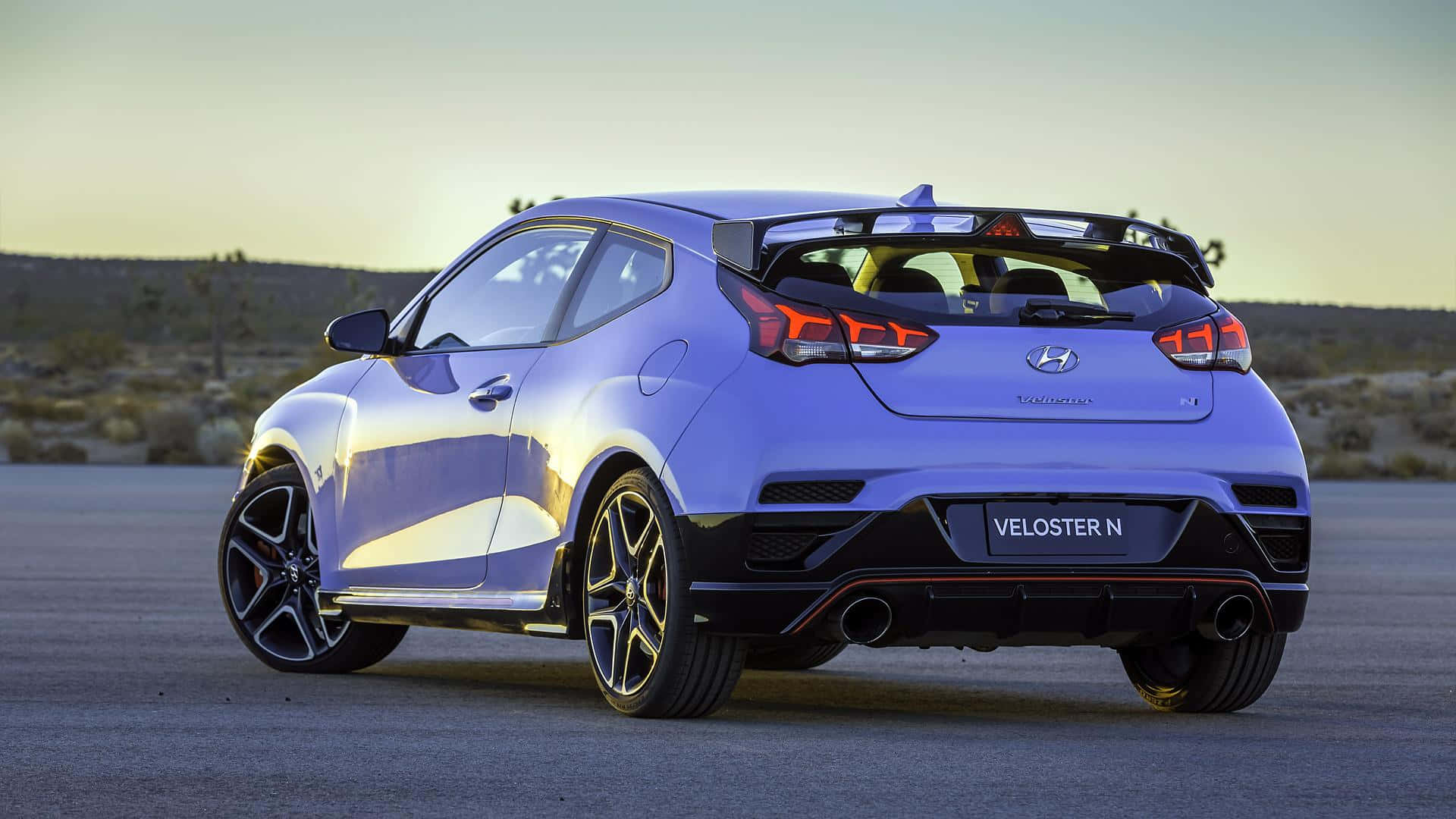 Hyundai Veloster 桌布 壁紙 — 一輛充滿活力的藍色現代Veloster N跑車停在沙漠公路上，背景是崎嶇的風景，正值黃金時刻。