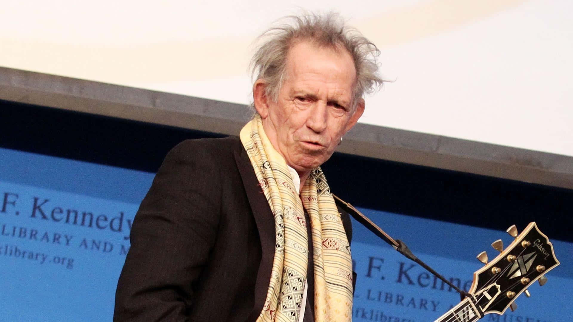  壁紙 — 一位頭髮灰白蓬鬆的老年男子，戴著圖案圍巾和黑色外套，在背景為 'F Kennedy Library' 的講台上拿著吉他。