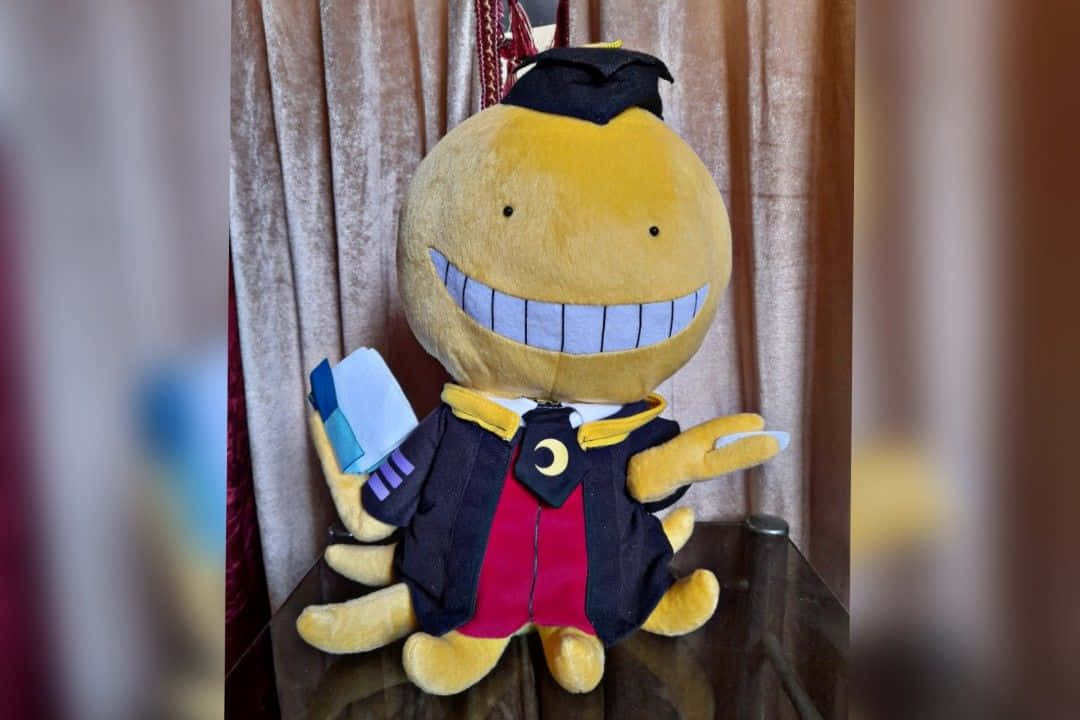 Koro Sensei 桌布 壁紙 — 一個大型黃色毛絨玩具，露出寬闊的牙齒笑容，戴著畢業帽和畢業袍，手持文憑。