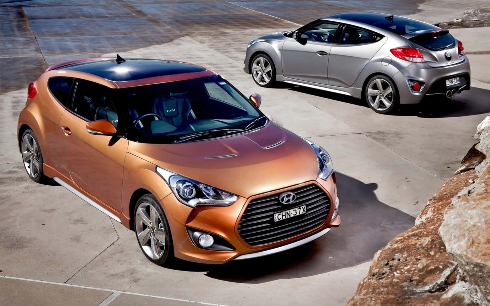 Hyundai Veloster 桌布 壁紙 — 兩輛時尚的現代Veloster跑車，一輛銅色一輛銀色，在明亮的日光下停在岩石懸崖旁的混凝土上。