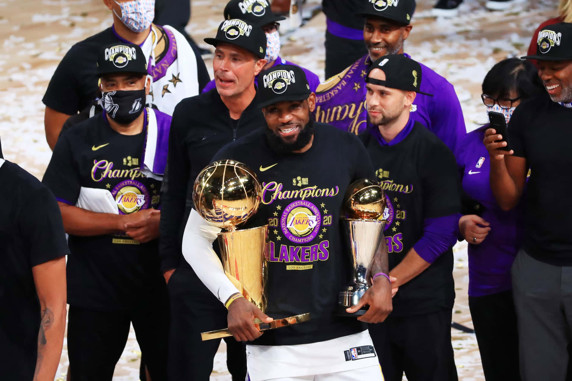  壁紙 — 勒布朗詹姆斯笑容滿面，手持兩座閃閃發光的NBA總冠軍獎盃，被身穿紫金色球衣的湖人隊友圍繞慶祝。