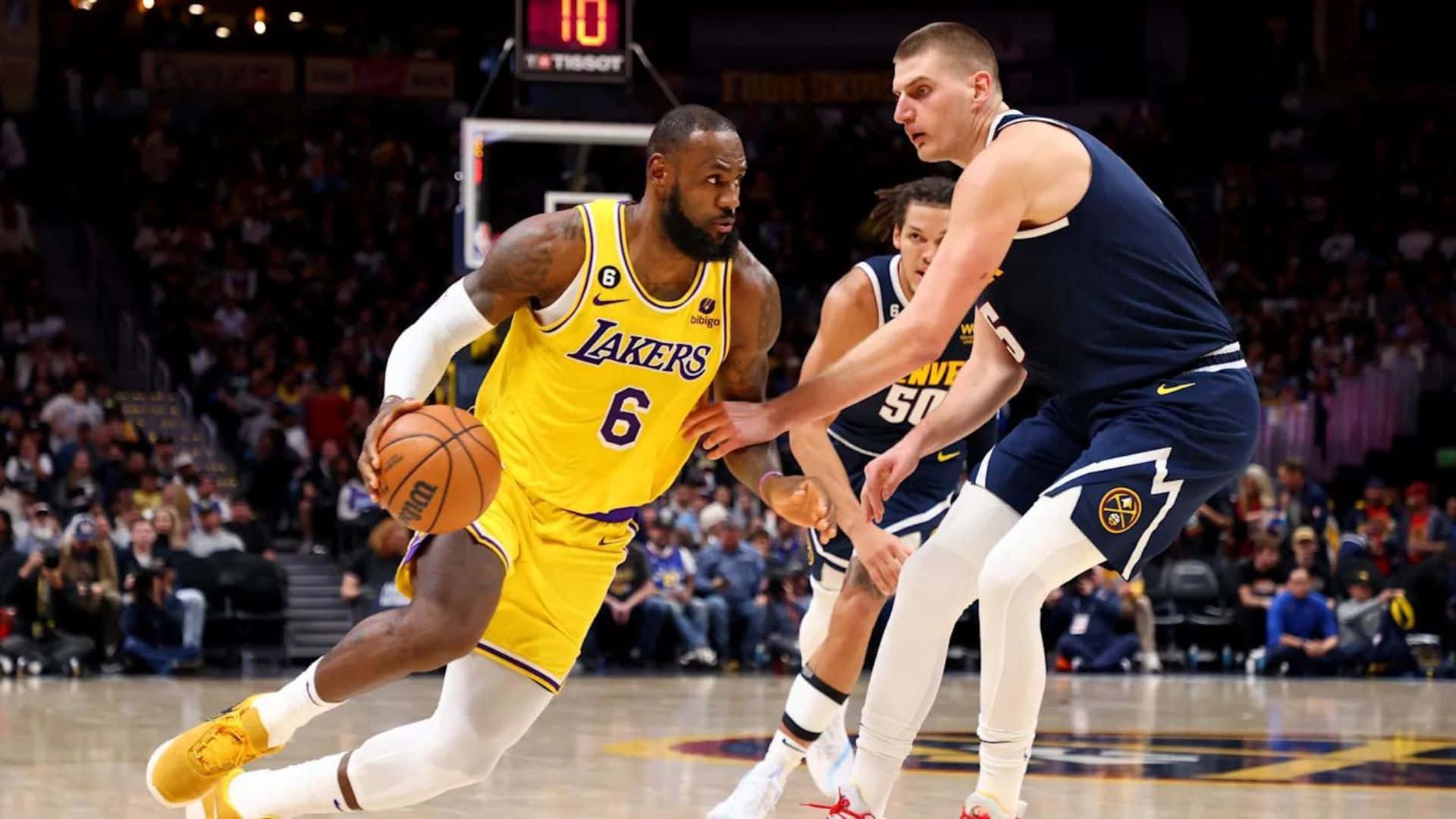 NBA Game 桌布 壁紙 — 勒布朗·詹姆斯身穿黃色湖人球衣，在場館燈光下於一場激烈的NBA比賽中運球越過丹佛金塊防守球員。
