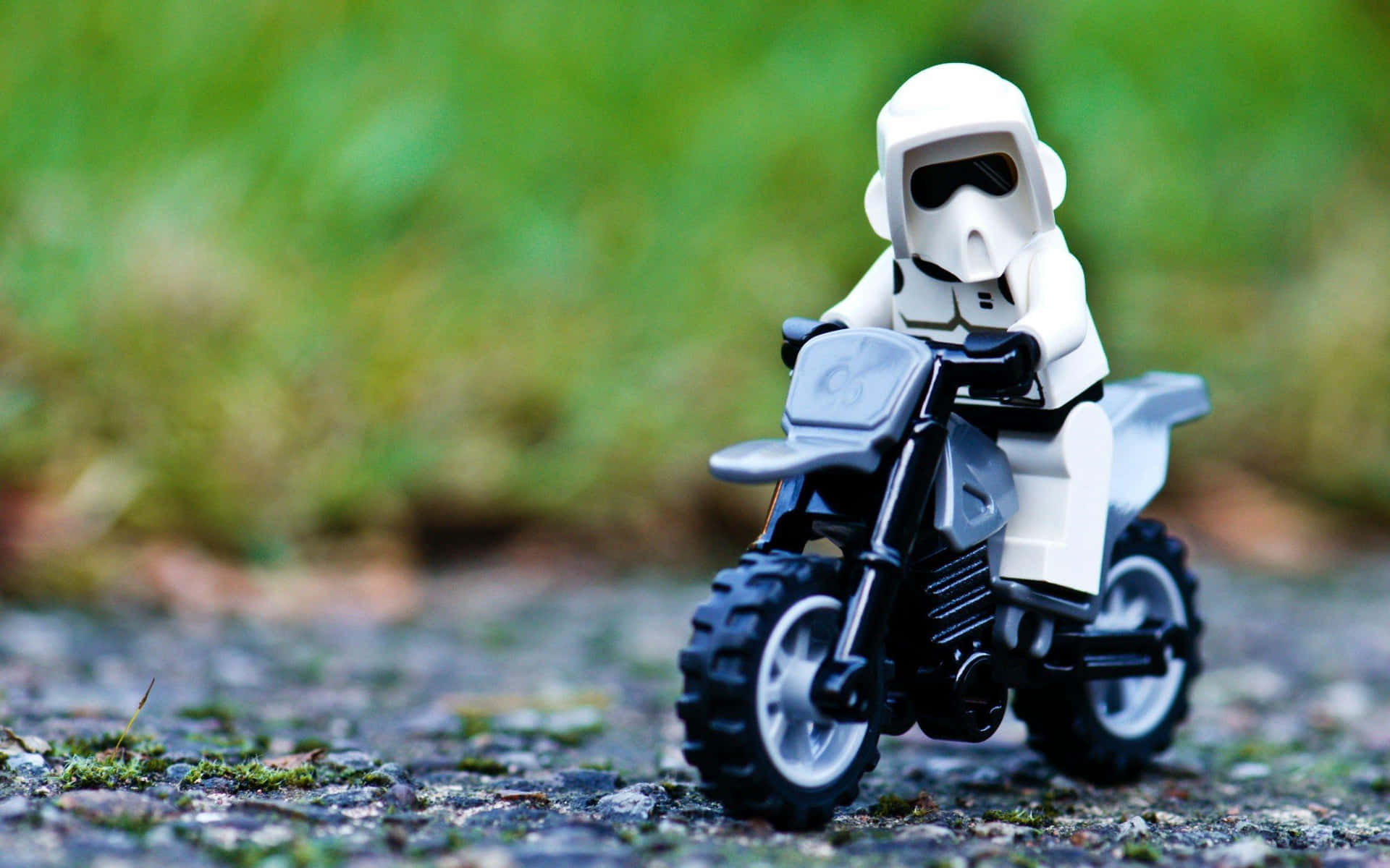  壁紙 — 一個白色的風暴兵LEGO迷你人偶騎著黑色和灰色的越野摩托車，在長滿青苔的鵝卵石小路上行駛，背景是模糊的綠色草地。