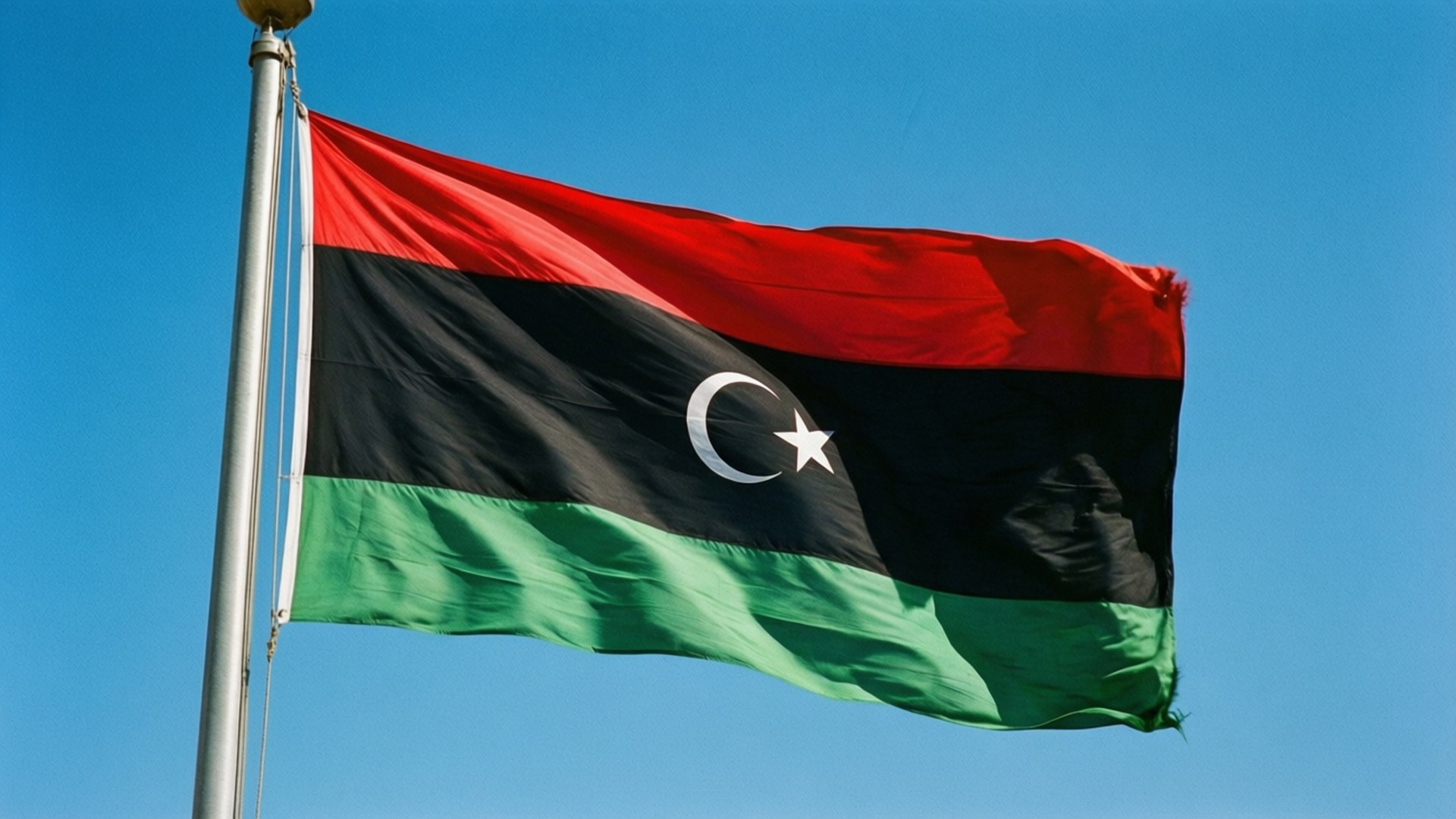 Libyan Flag Flying 壁紙 — 利比亞國旗，帶有紅、黑、綠條紋和白色新月及星星，在清澈的藍天下自豪地飄揚。