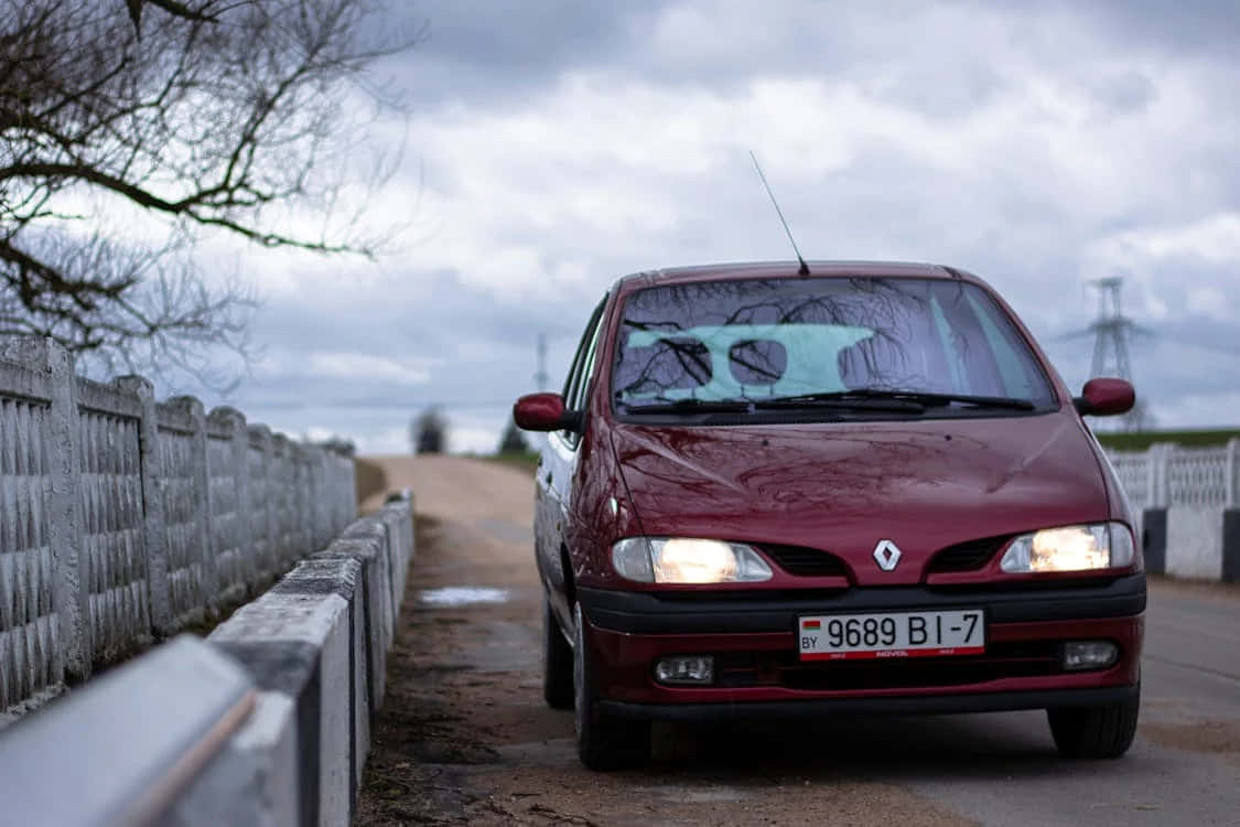 Renault Scenic 桌布 壁紙 — 一輛栗色雷諾汽車亮著頭燈，停在鄉村道路上，旁邊是風化的混凝土護欄，天空多雲。