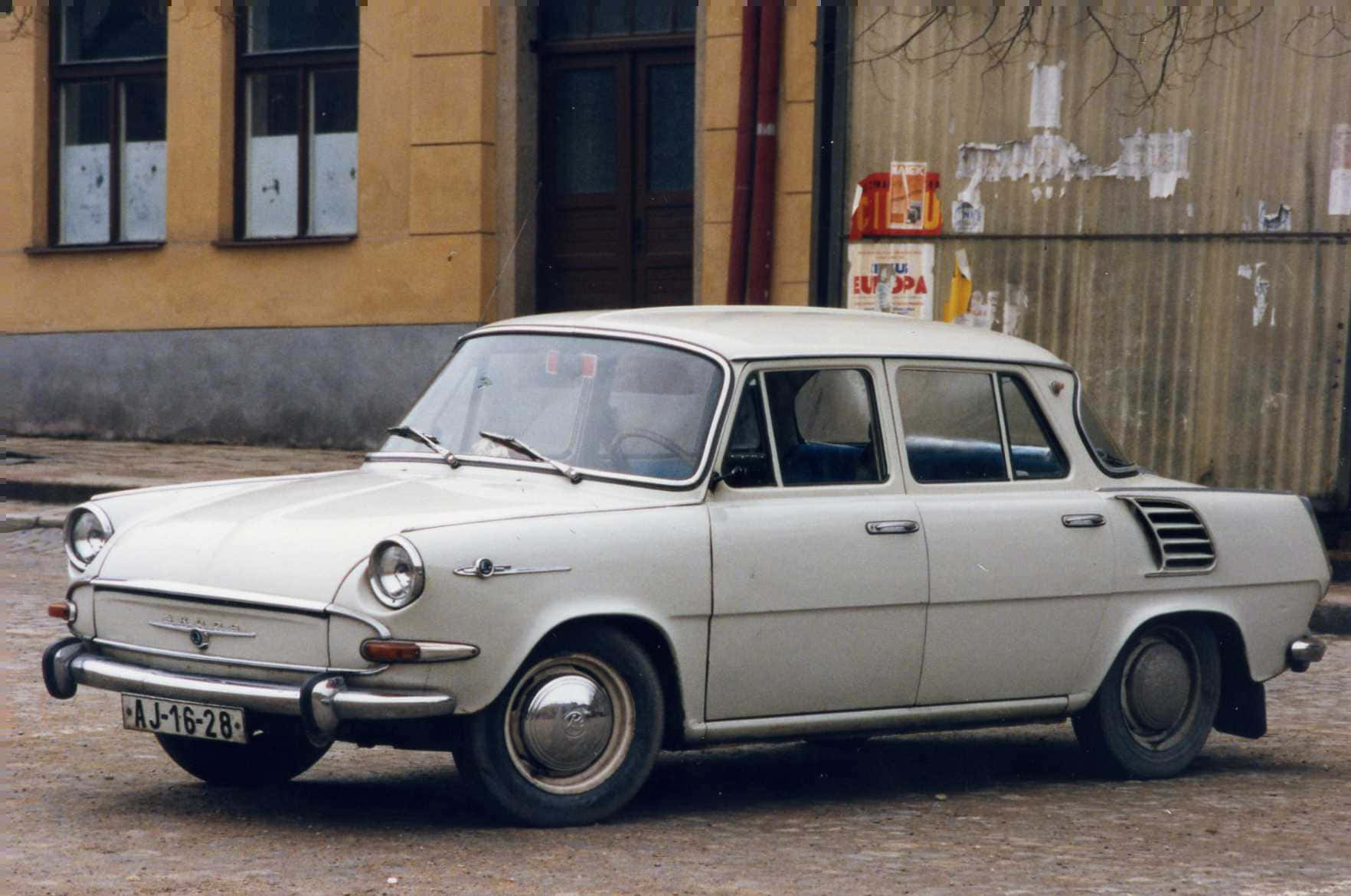  壁紙 — 一輛白色復古Škoda 1000 MB轎車停在鵝卵石街道上，旁邊是一座風化的黃色建築。