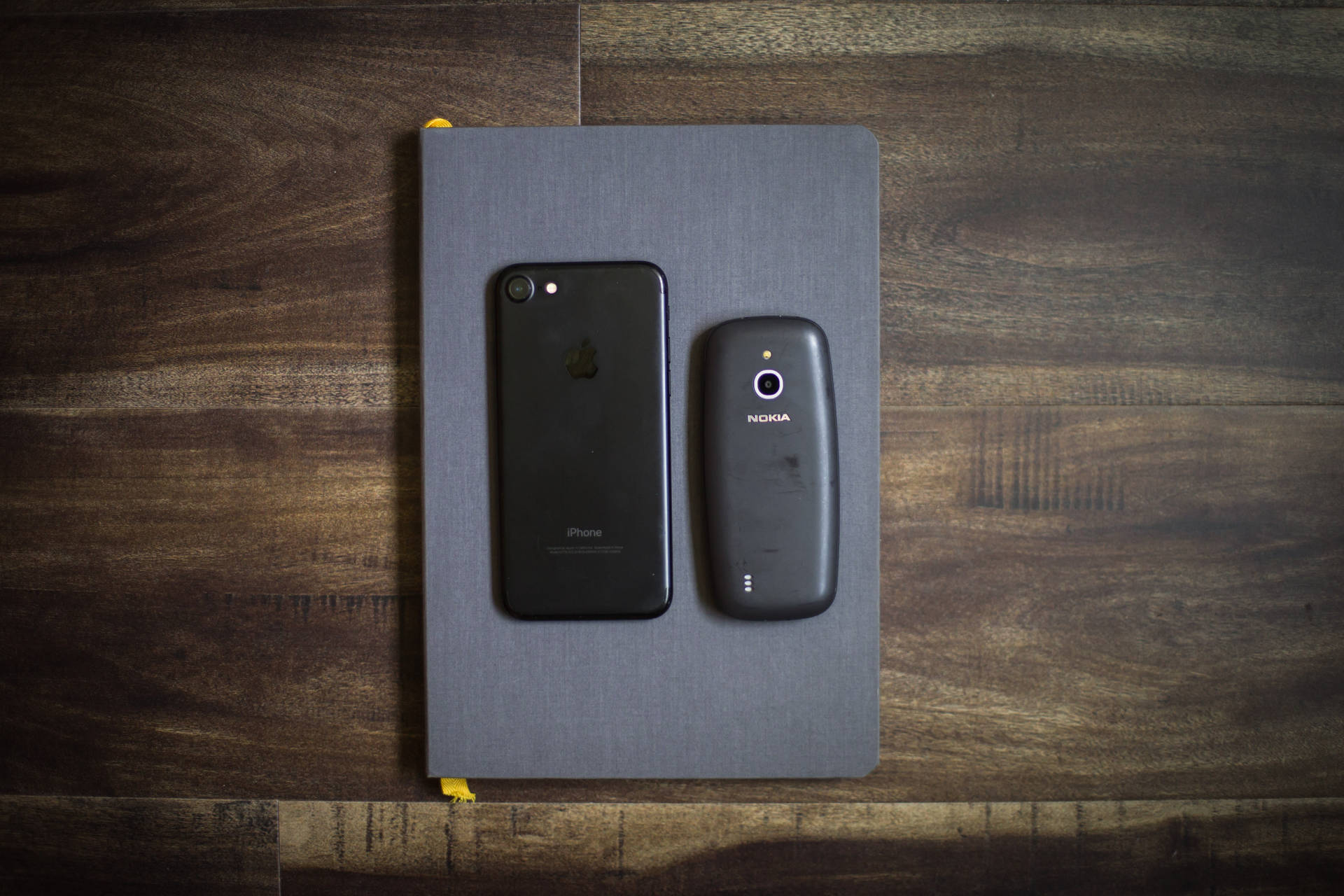 木頭上灰色筆記本上的兩部舊智慧型手機iPhone和Nokia。 — iPhone 11 壁紙