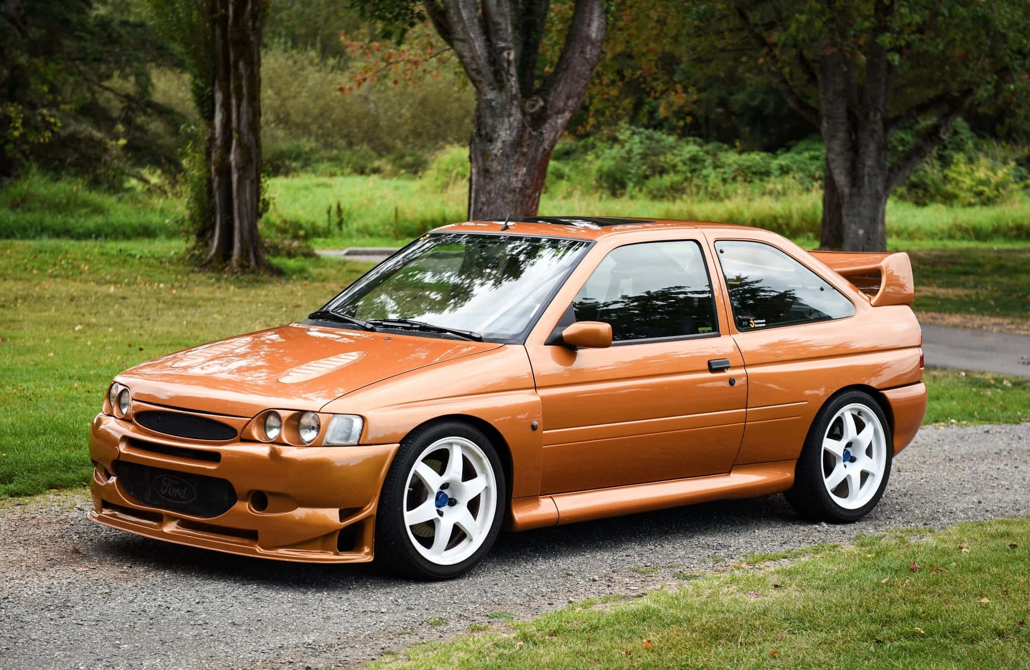 Ford Escort 桌布 壁紙 — 一輛亮橙色的福特 Escort RS Cosworth 跑車停放在草地和樹木旁邊的碎石上。