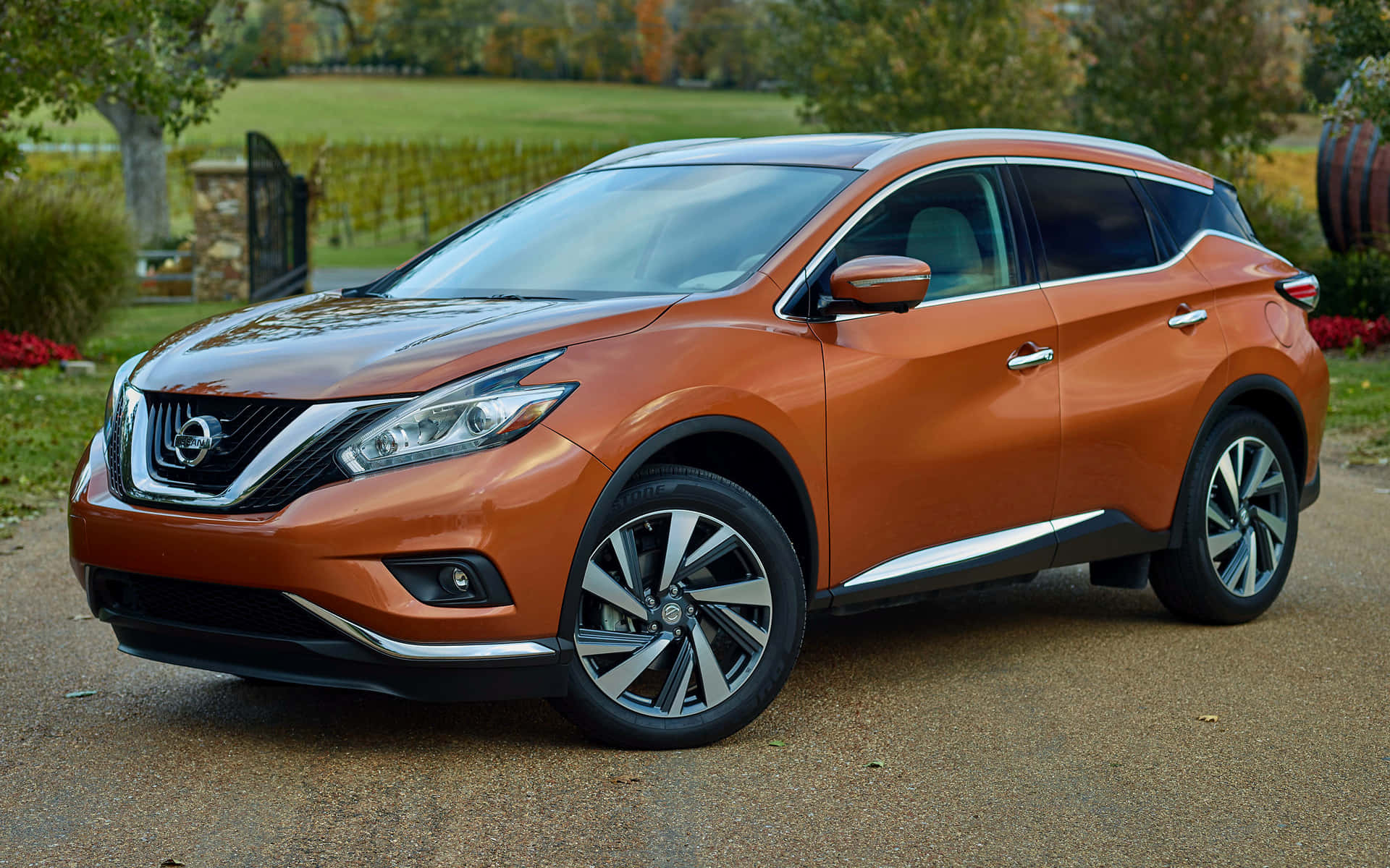 Nissan Murano 桌布 壁紙 — 一輛亮橙色日產Murano SUV停泊在碎石車道上，背景有秋天的樹木和石籬笆。
