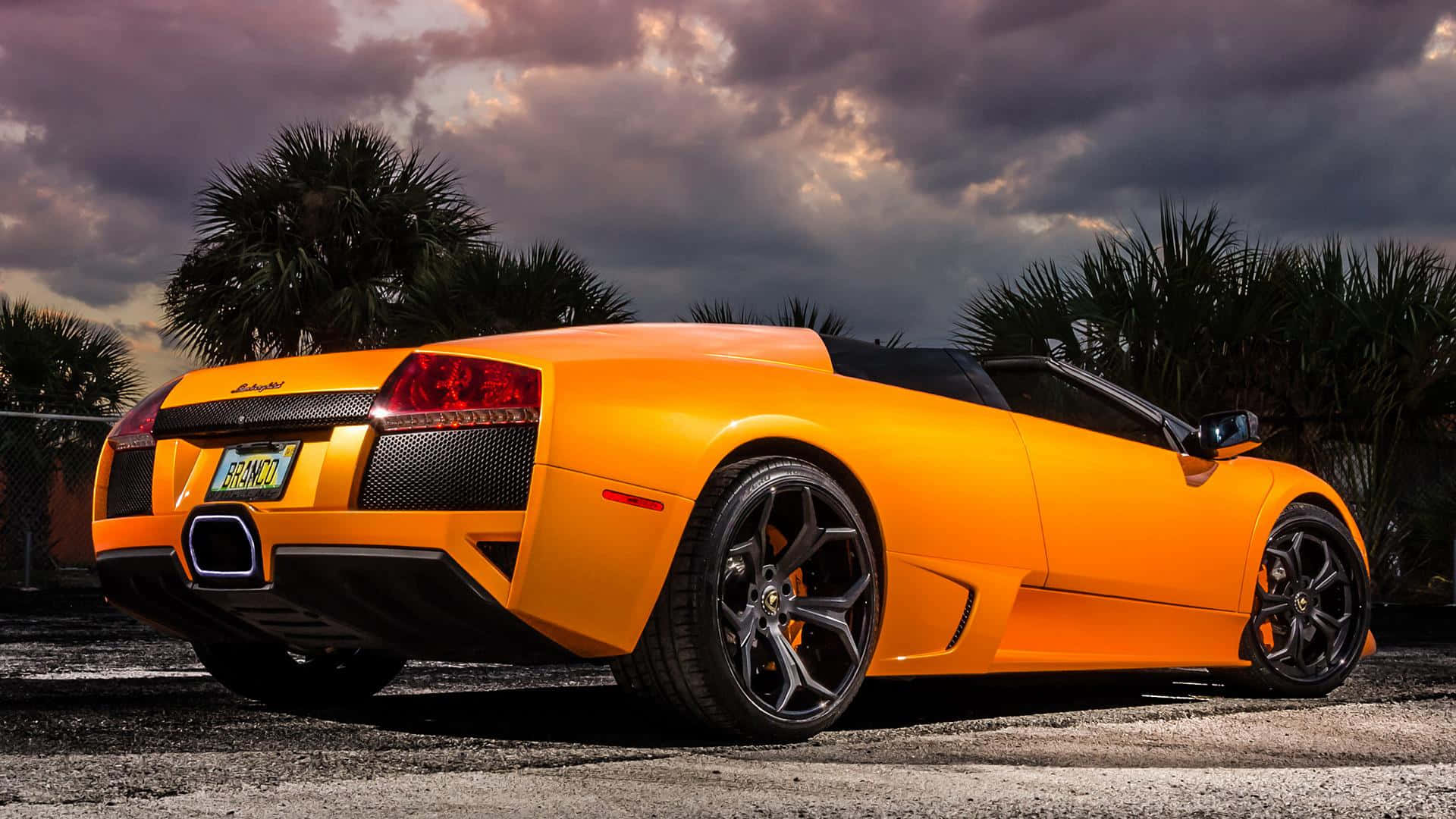 Lamborghini Murciélago 桌布 壁紙 — 一輛充滿活力的橙色藍寶堅尼 Murciélago Roadster 停泊在瀝青路上，背景是戲劇性的暮色天空和棕櫚樹。