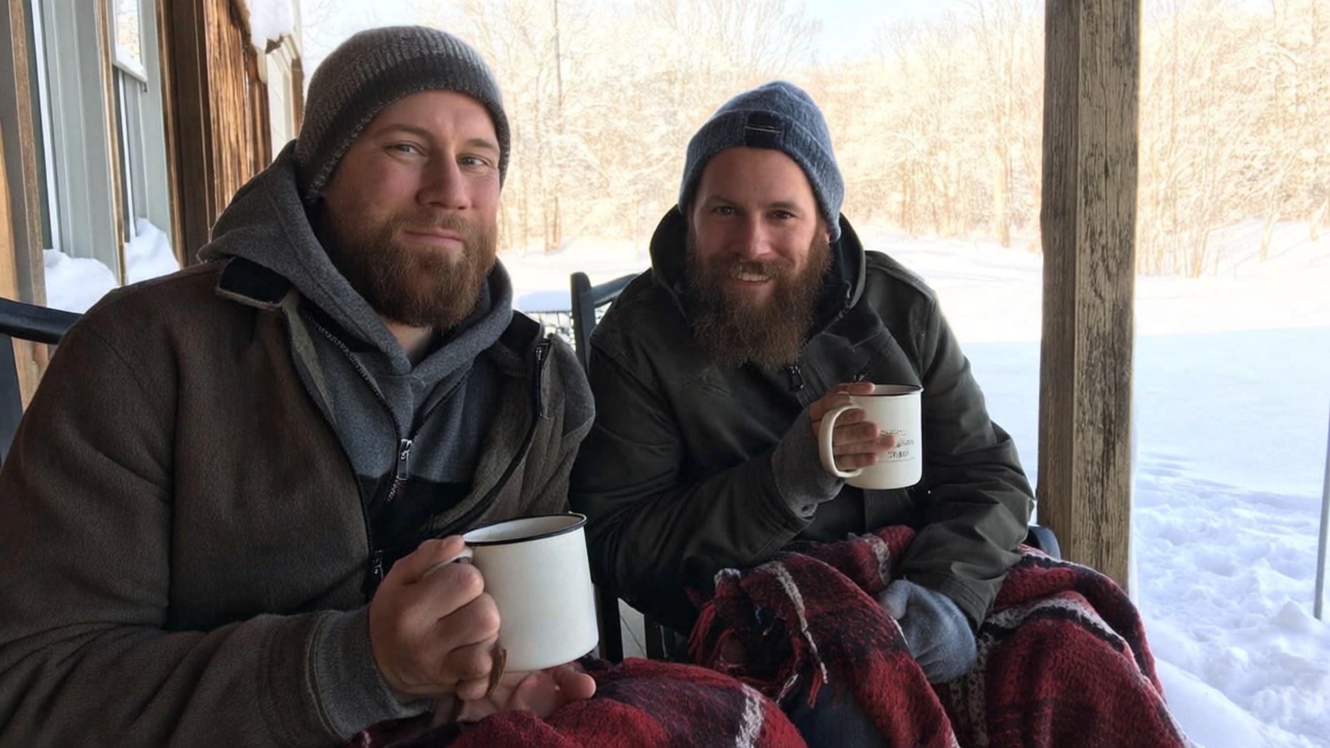 Cozy Winter Morning 壁紙 — 兩個留鬍鬚的男人戴著毛帽，在積雪的門廊上裹著格紋毯，從杯子裡啜飲熱飲。
