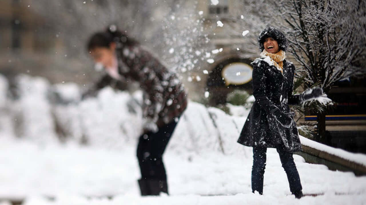  壁紙 — 兩個人在白雪覆蓋的市區公園裡快樂地玩耍，雪花在他們周圍飛舞。