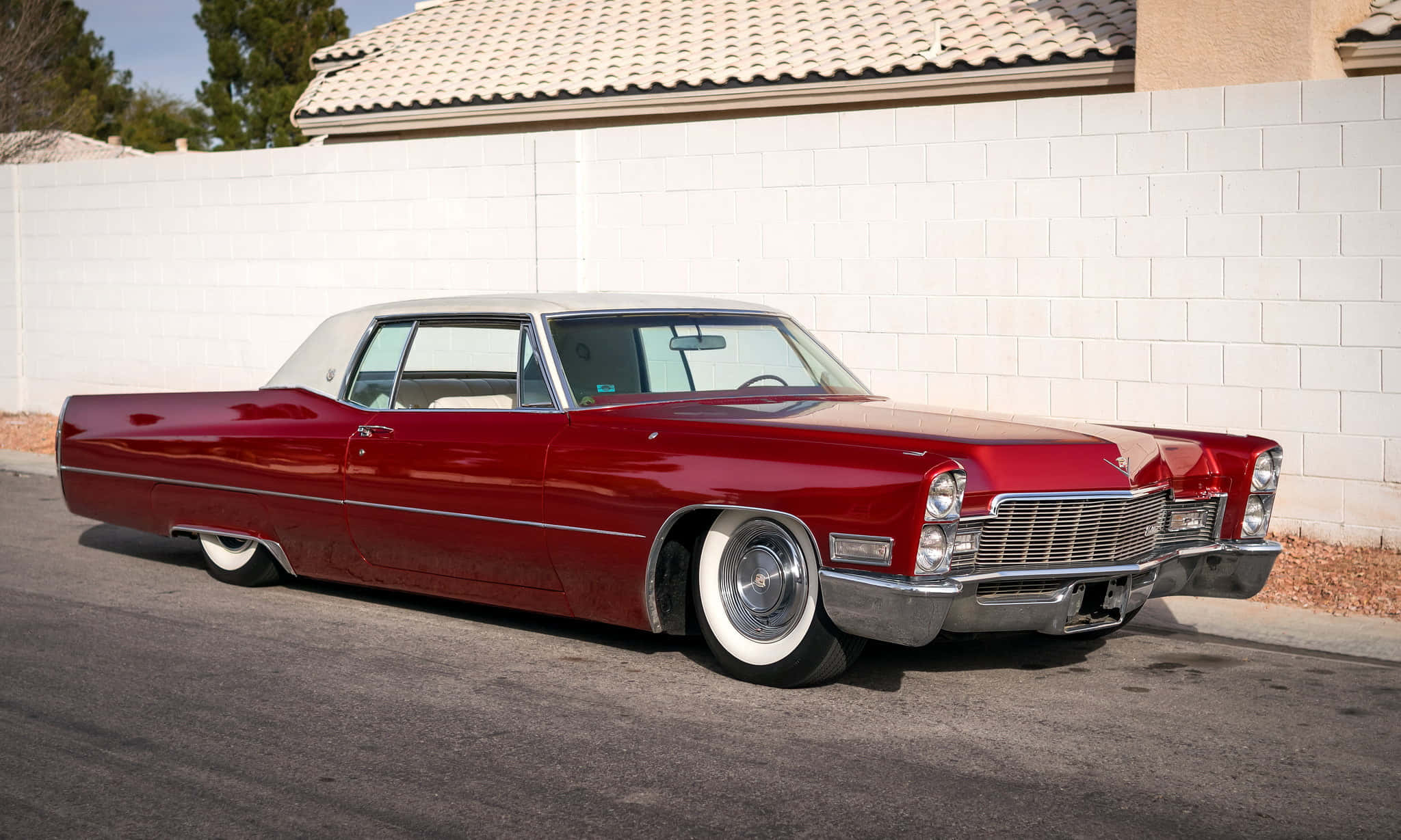 Cadillac Deville 桌布 壁紙 — 一輛光澤的紅色經典凱迪拉克，配備白色側壁輪胎，停在瀝青路面上，旁邊是白色磚牆，在明亮的日光下。