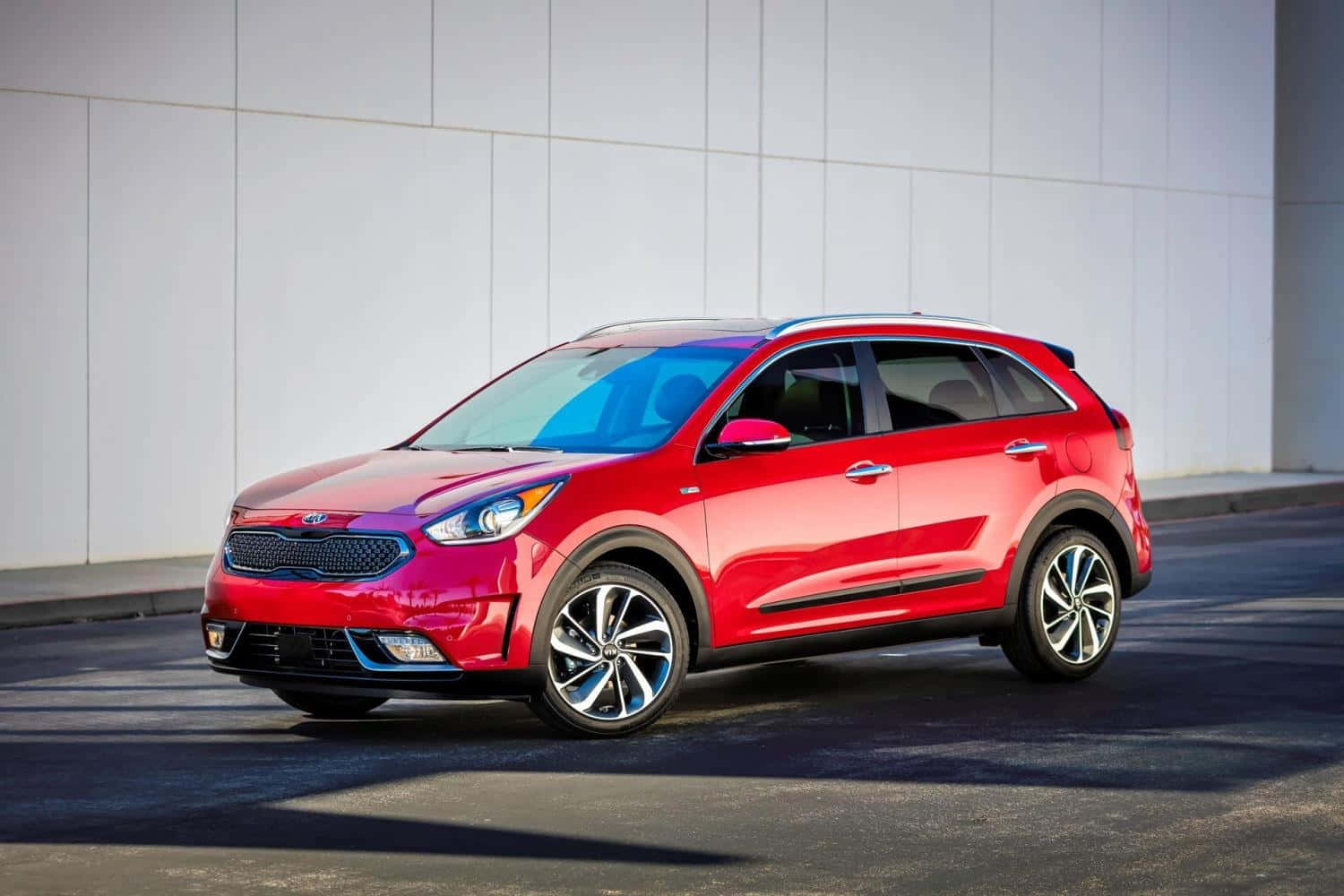 Kia Niro 桌布 壁紙 — 一輛光澤的紅色Kia SUV停在瀝青路上，明亮的陽光在純白牆壁上投下銳利的陰影。